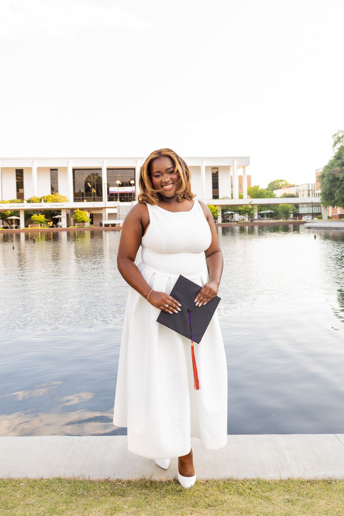Proud Black graduate in white dress holds diploma cover while standing by a reflective campus pond.