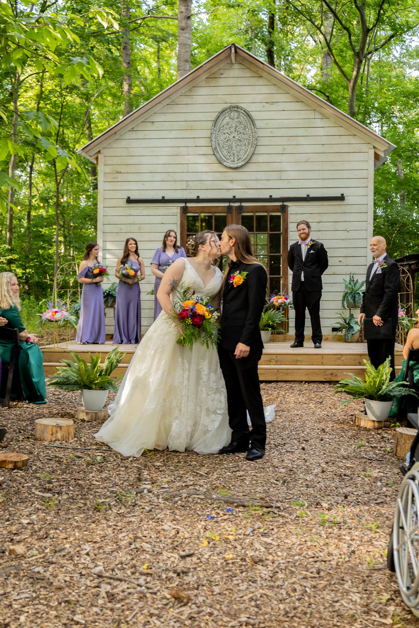 Two brides share first kiss at outdoor woodland wedding ceremony with bridesmaids in lavender dresses before white chapel.