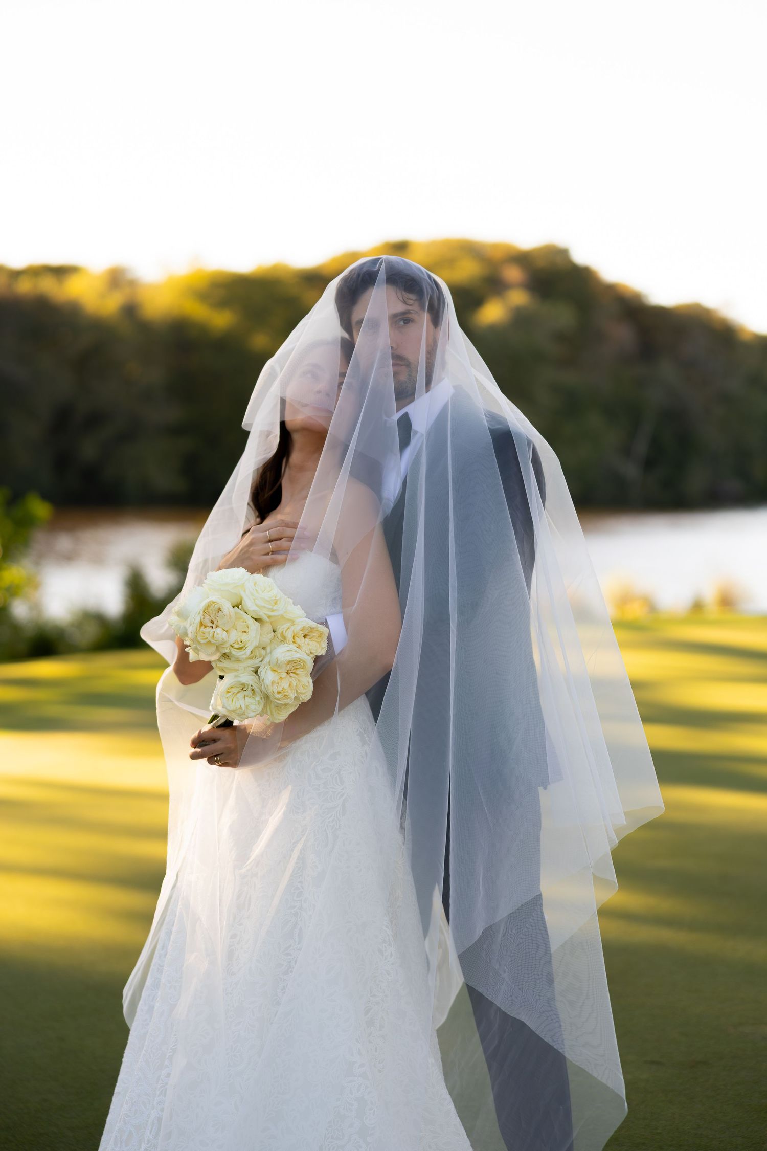 Bride and groom sharing a kiss under a white veil outdoors, bride holding white rose bouquet at golden hour.