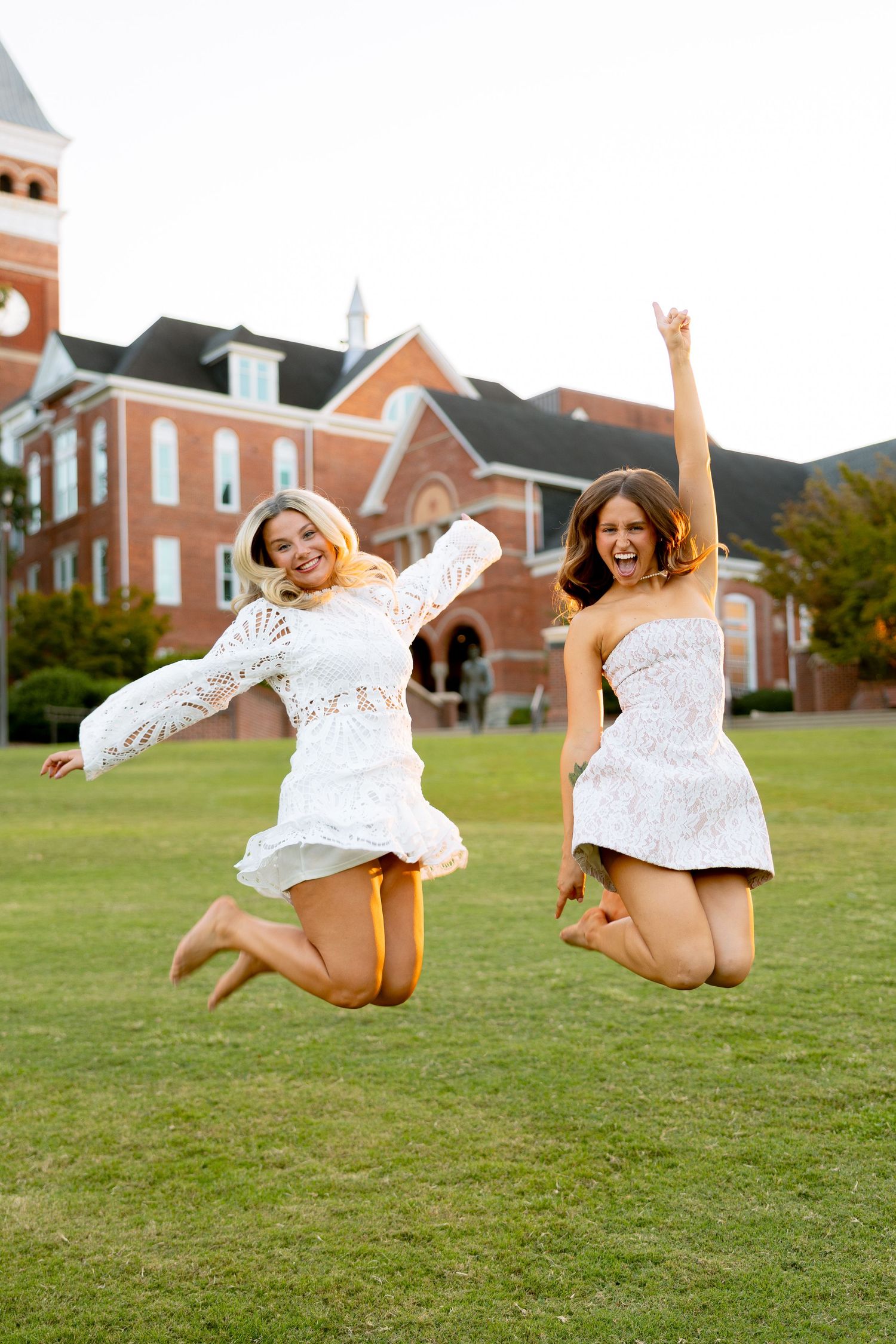 Two women in white dresses jumping joyfully on a green lawn in front of a red brick building.
