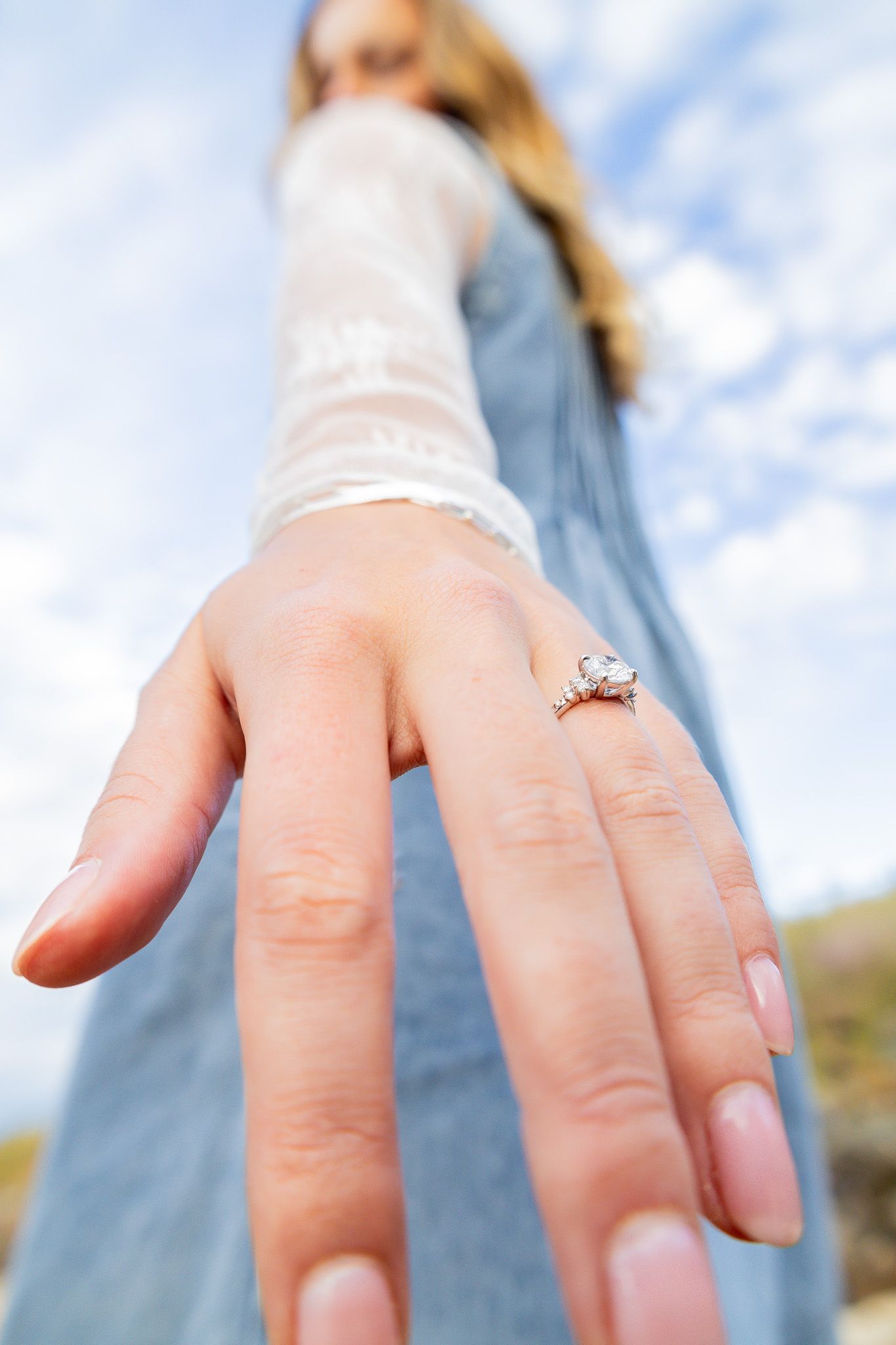 Close-up shot of a diamond engagement ring on a hand with a blurred outdoor background.