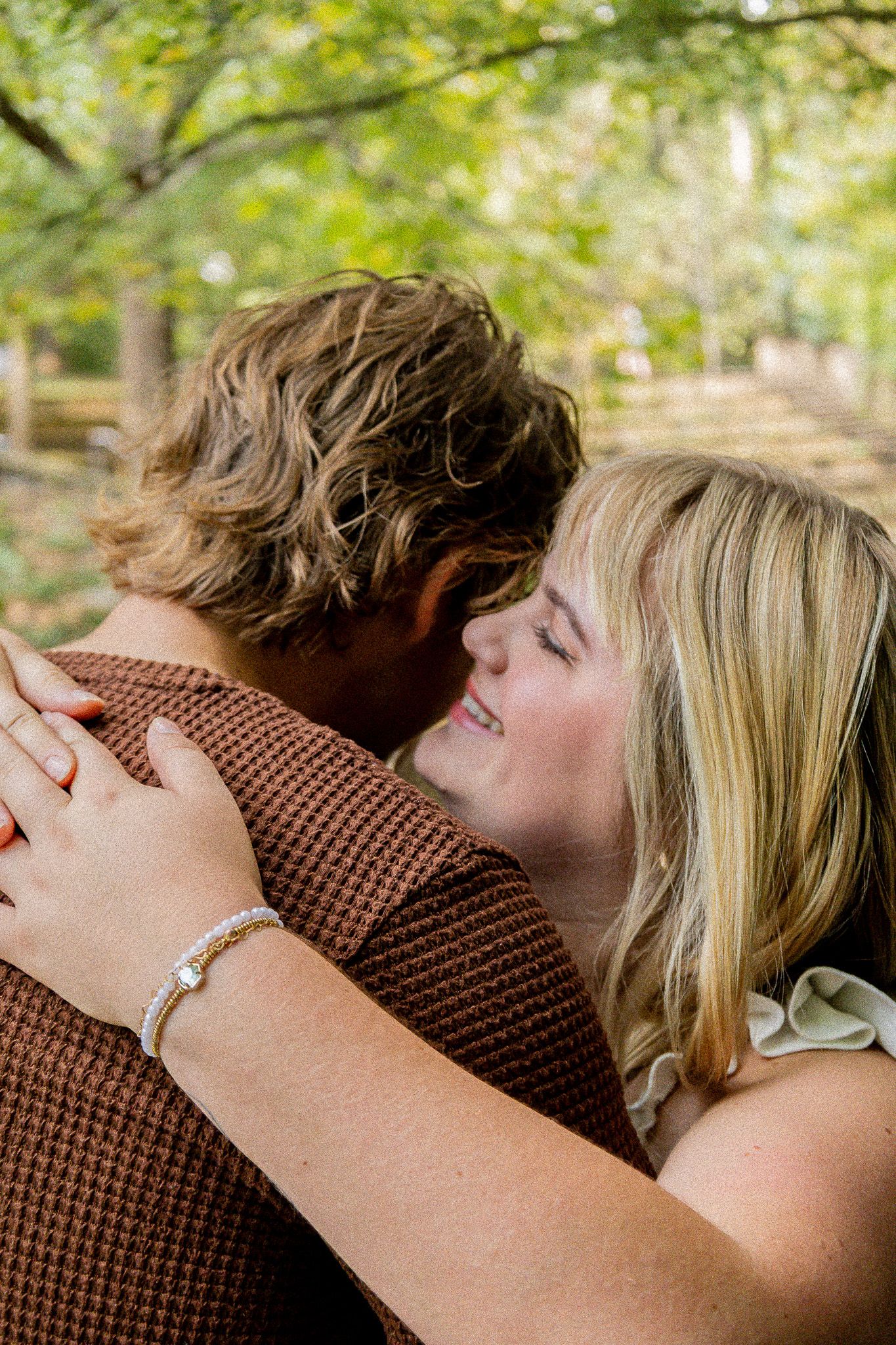 A couple embraces outdoors, woman with blonde hair and bracelet smiling as man in brown shirt holds her among green trees.