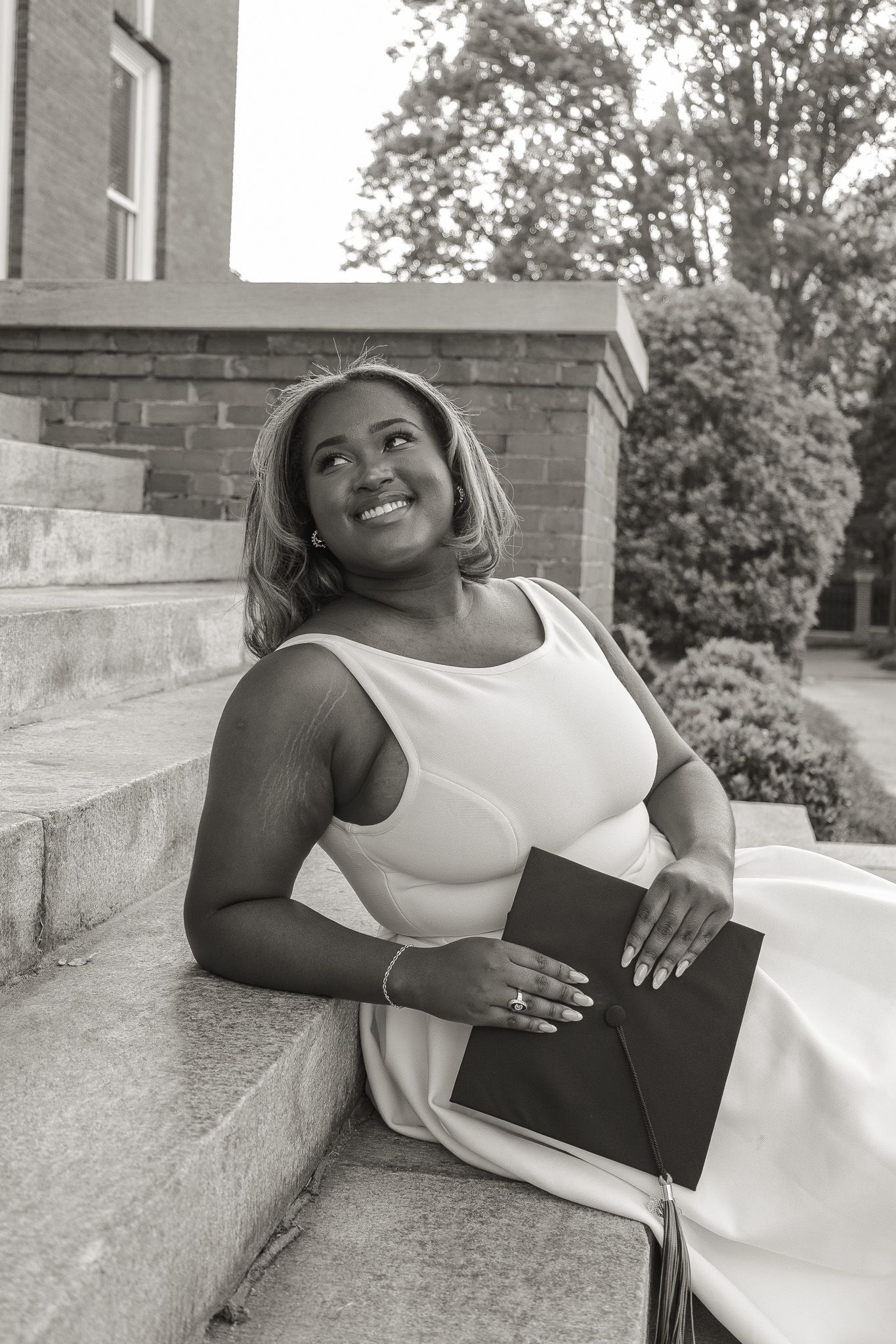 Smiling graduate in white dress holds diploma on stone steps outside brick university building, black and white photo.