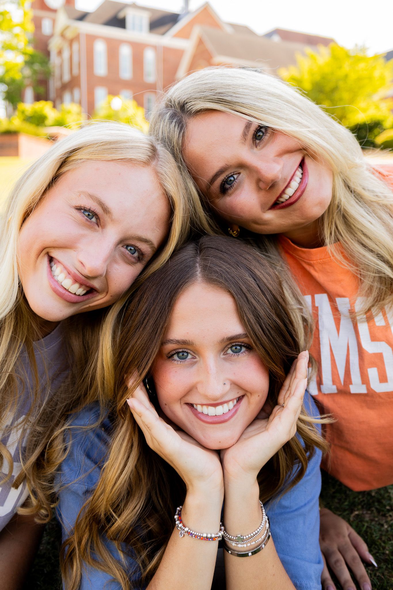 Three smiling young women pose together outdoors near a brick building, wearing casual college sweatshirts on a sunny day.