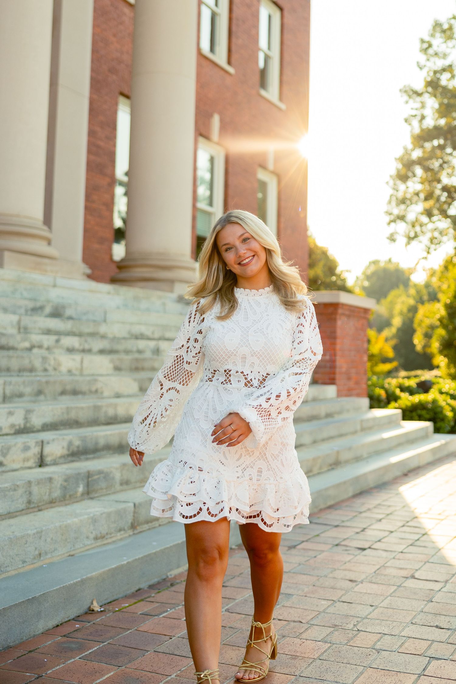 Young blonde woman in white lace dress poses on brick steps outside a historic building at golden hour.