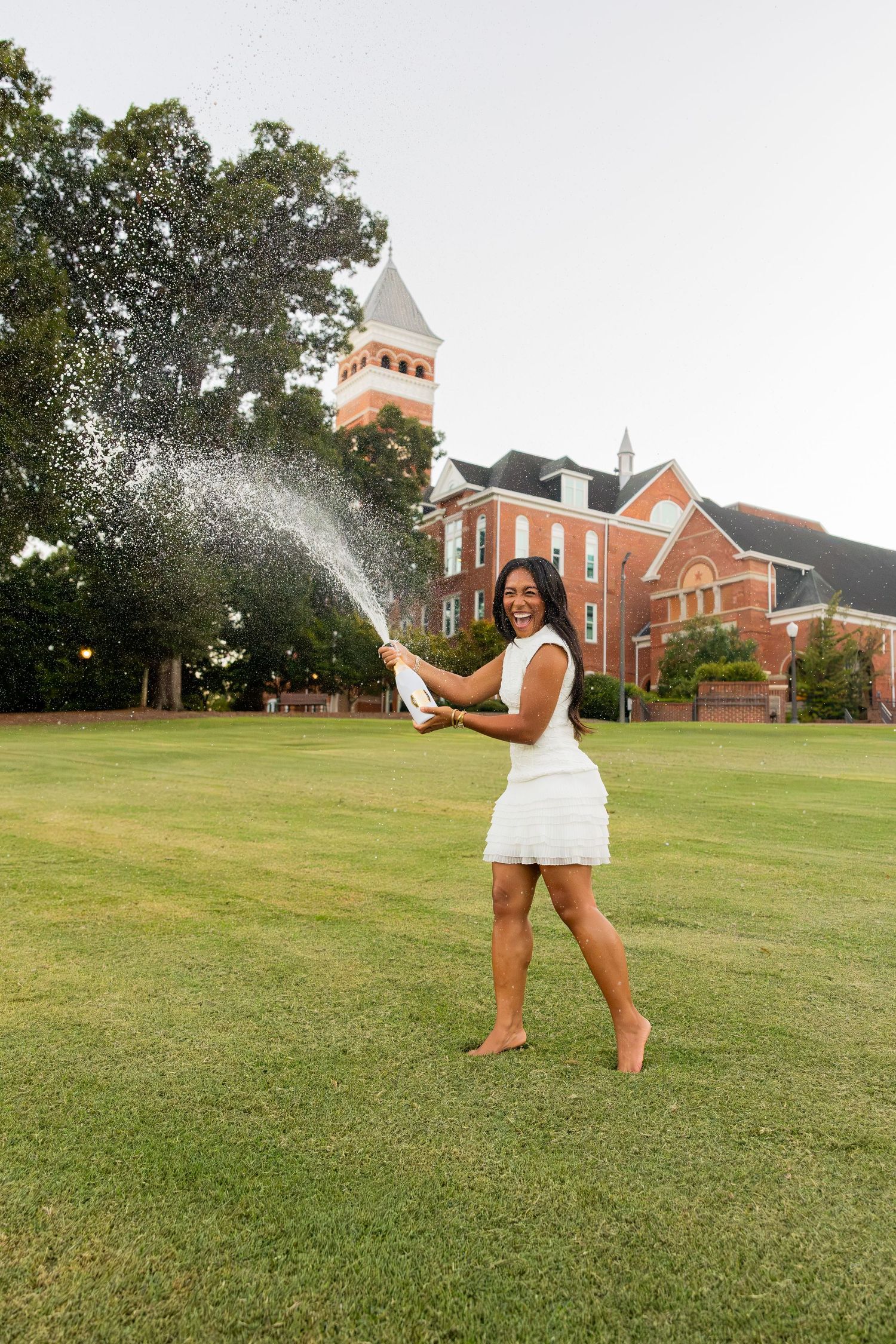 Young woman in white dress celebrates graduation by spraying champagne on campus lawn at dusk.