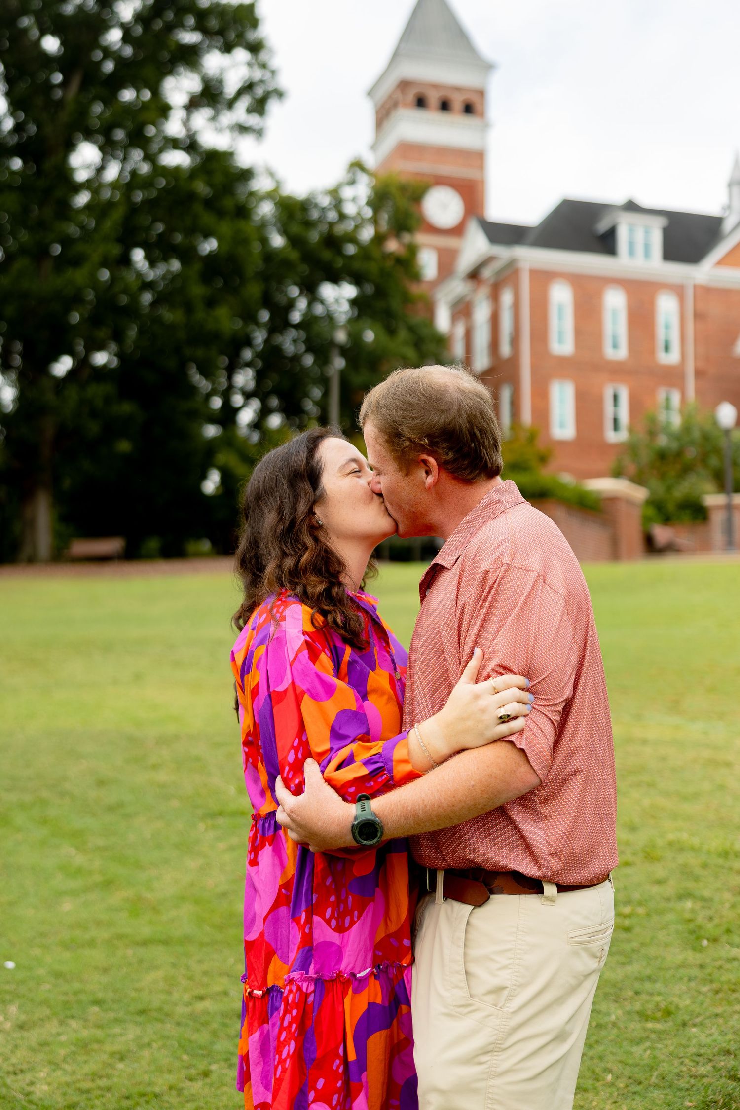 Couple sharing a romantic kiss on green lawn with historic red brick building and clock tower in background.
