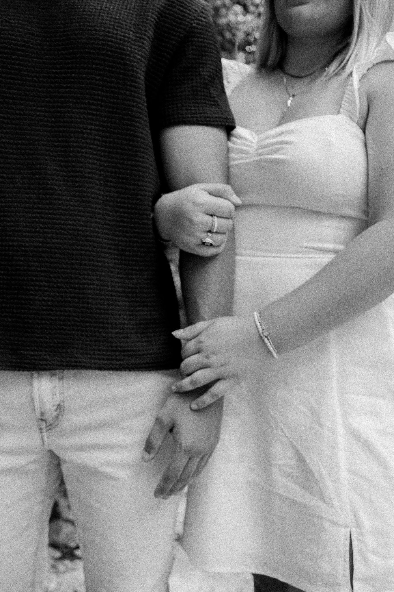 Black and white photo of a couple holding hands, woman in white dress with ring and bracelet, man in dark shirt.