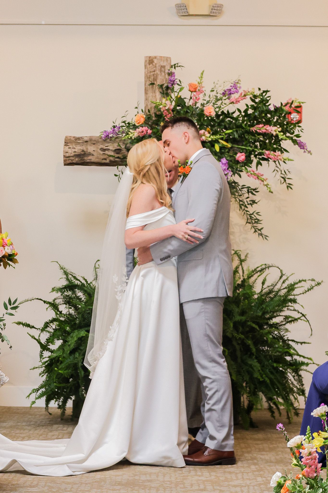 Bride and groom share first kiss at altar decorated with wooden cross and colorful floral arrangements.