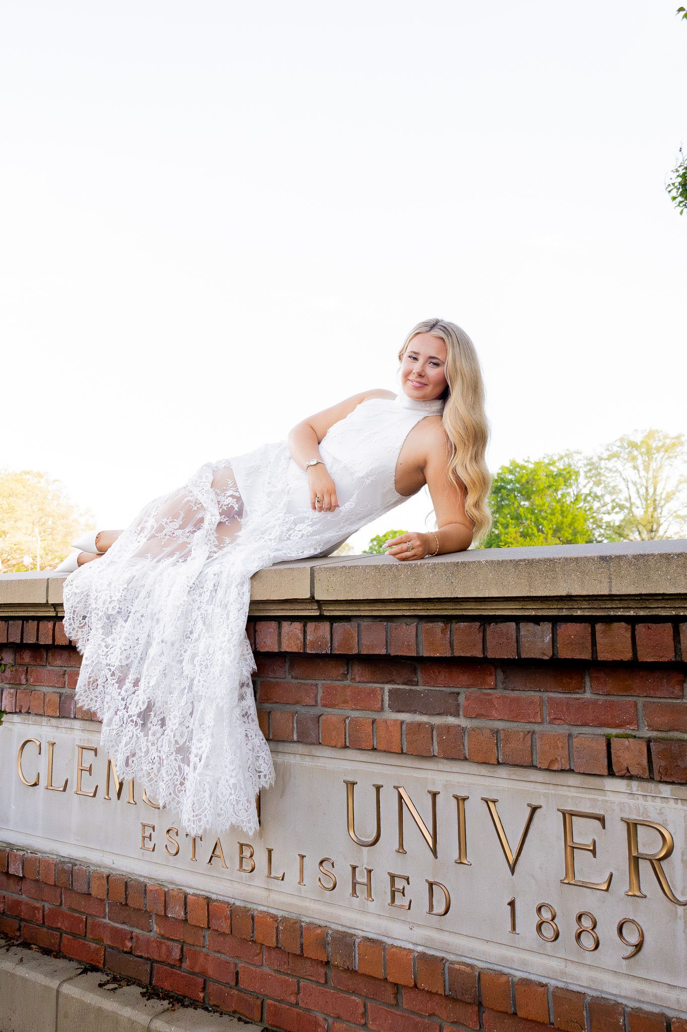 Blonde woman in white lace gown posing on Clemson University brick entrance wall, established 1889.