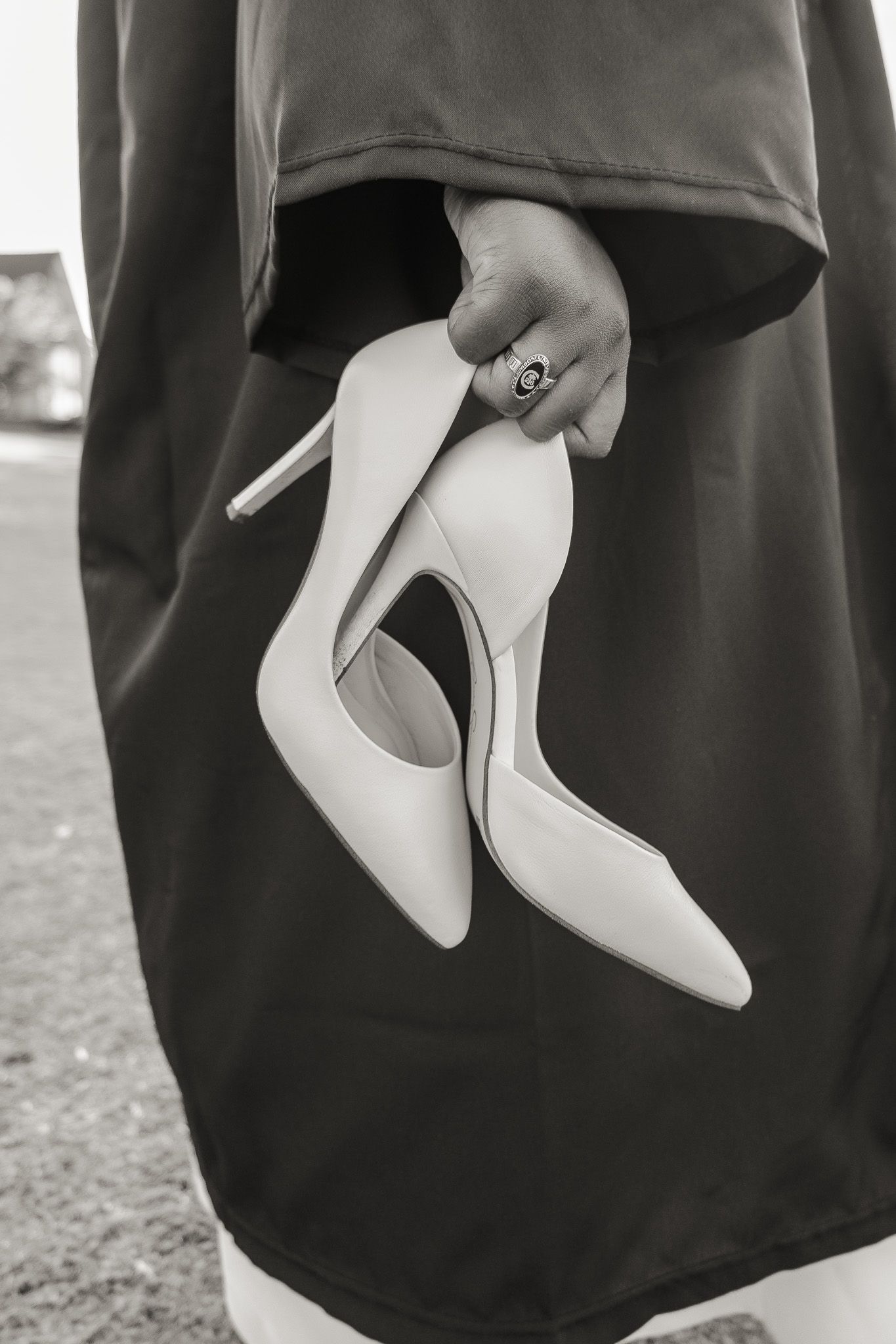 Black and white photo of a hand holding white high heel shoes against a dark graduation gown.