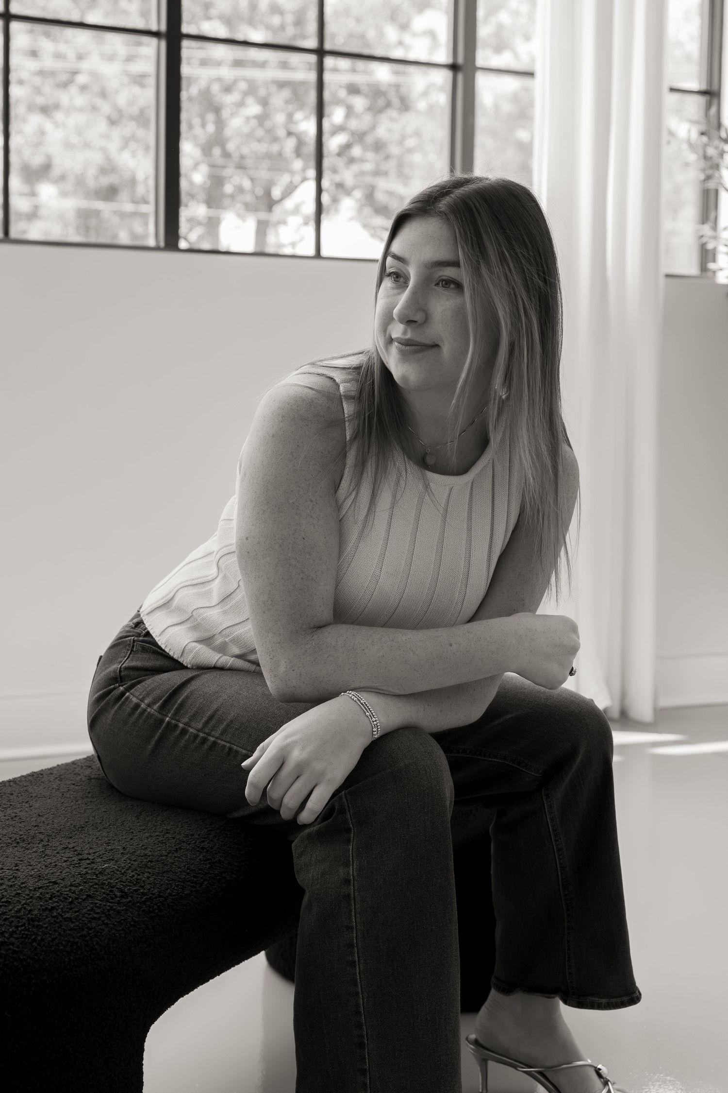 Black and white portrait of a young woman sitting on a bench, wearing jeans and a sleeveless top, gazing thoughtfully.