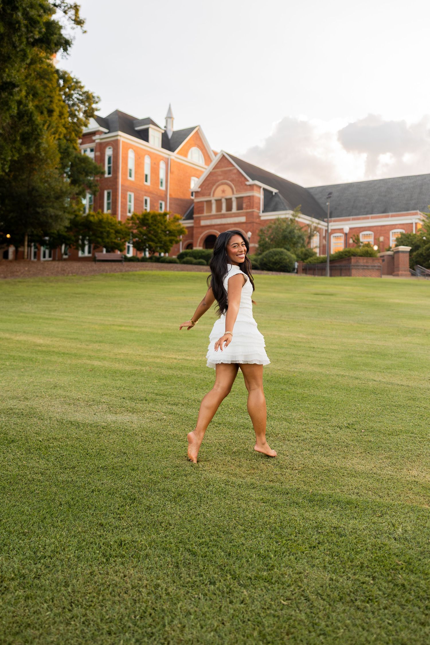 Joyful young woman in white dress walks barefoot across green lawn near brick university building.