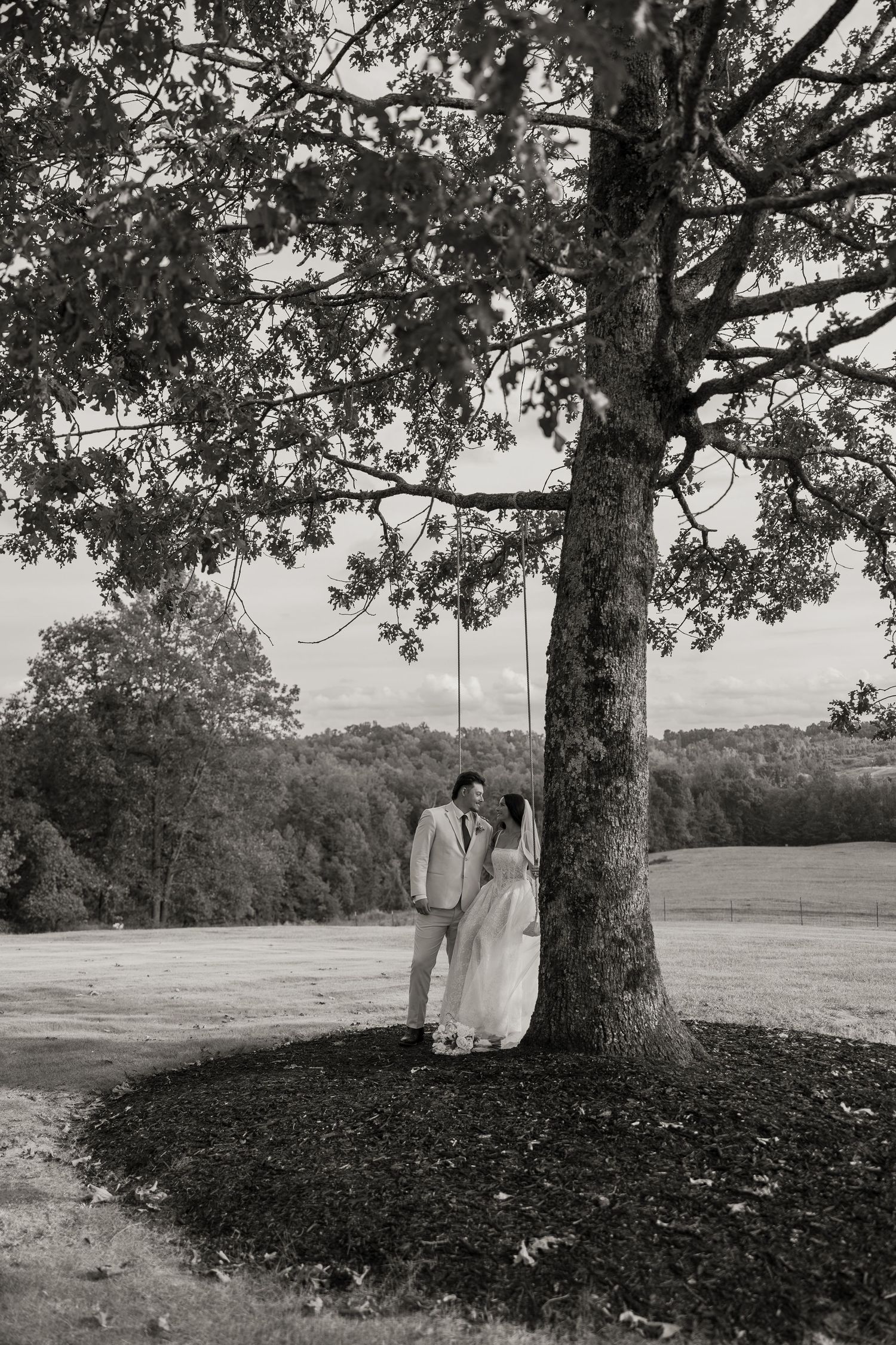 Black and white photo of a bride and groom embracing under a large tree with a rope swing in a scenic outdoor setting.