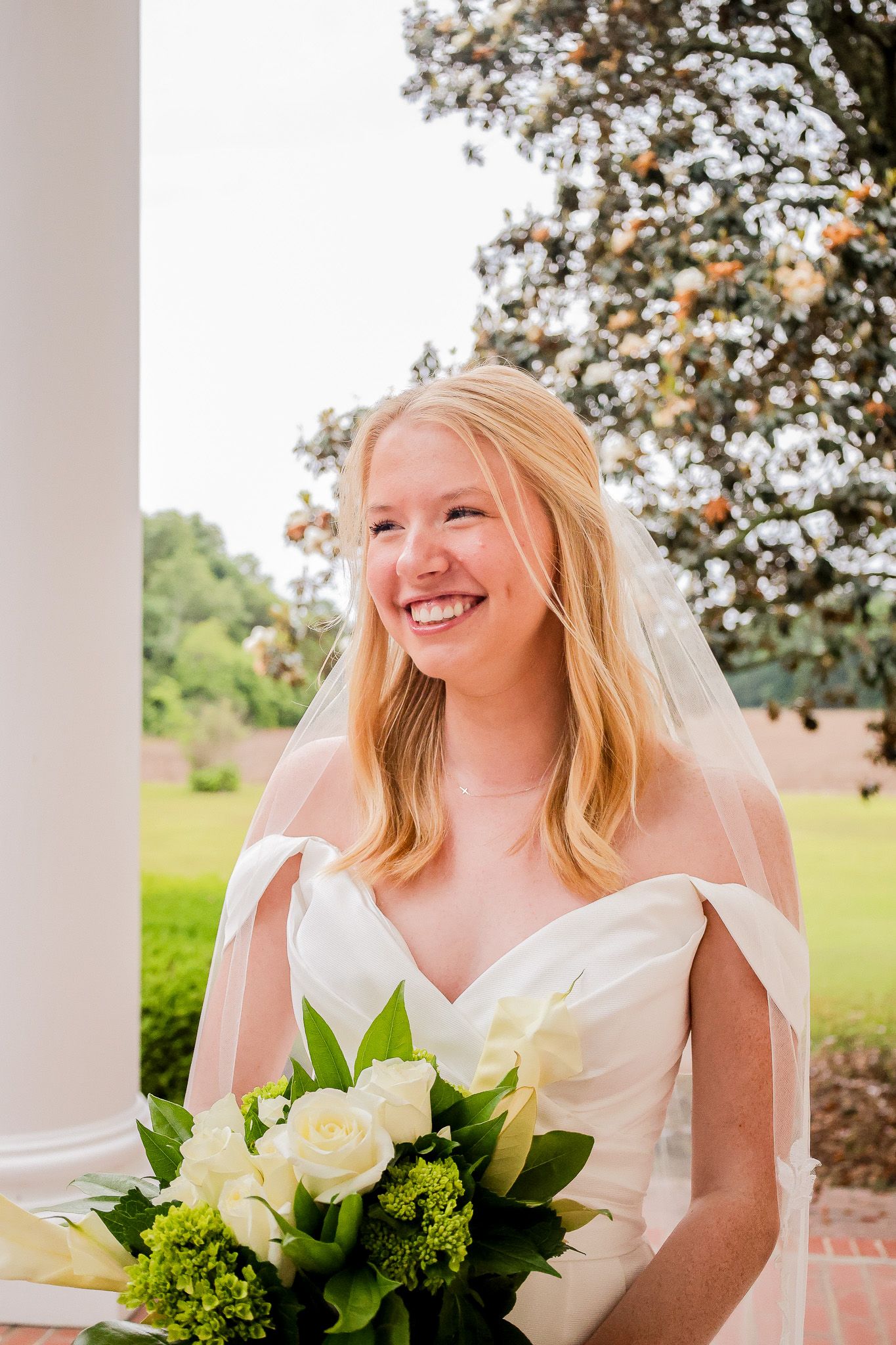 Smiling blonde bride in white gown and veil holds white roses bouquet outdoors near white column.