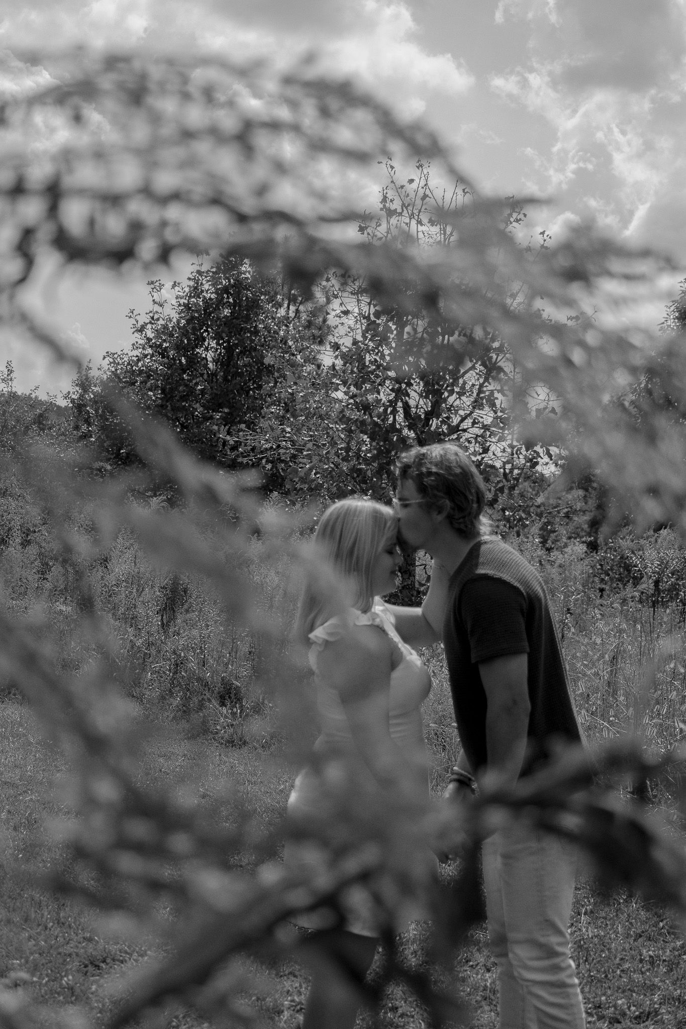 Black and white photo of a couple sharing a romantic kiss outdoors, framed by blurred branches with lush trees behind.