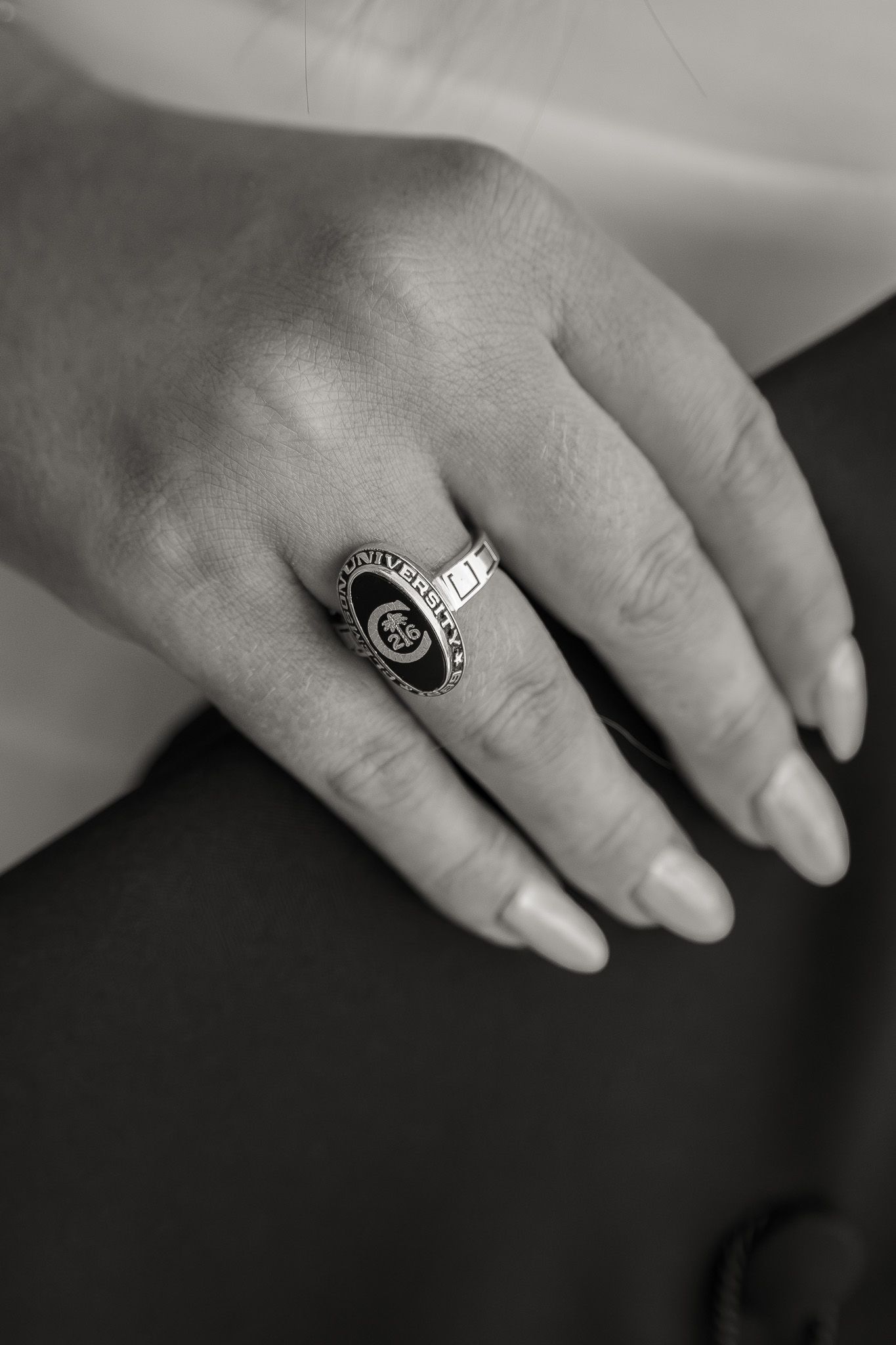 Close-up of a hand wearing a black and silver university class ring with manicured nails, in black and white.