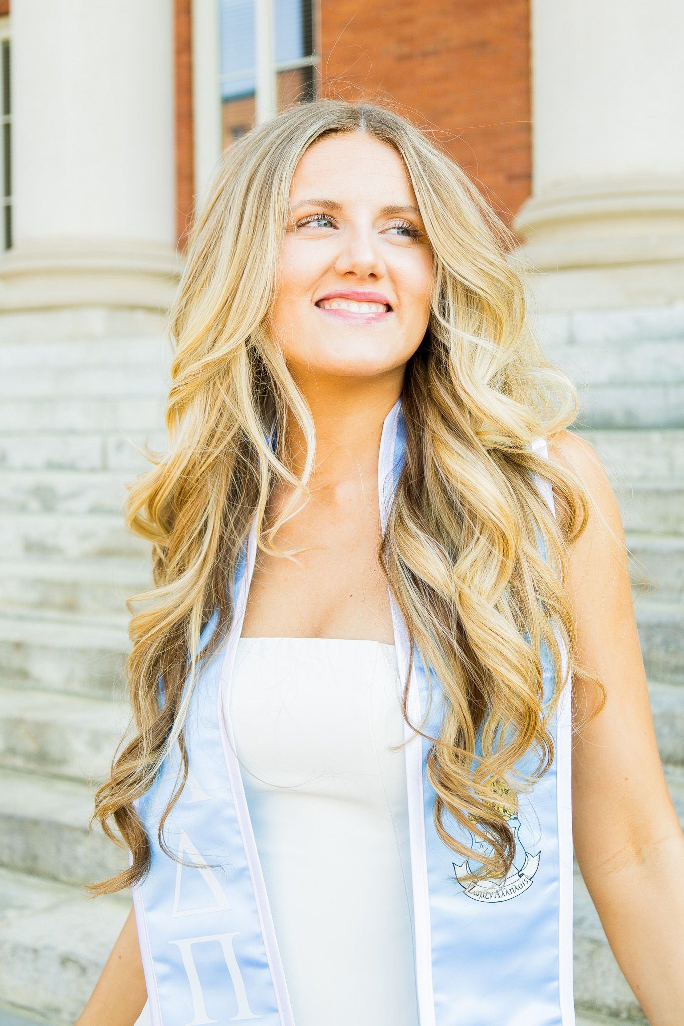 Smiling blonde female graduate wearing white dress and graduation stole posing on steps of a classic brick university building.