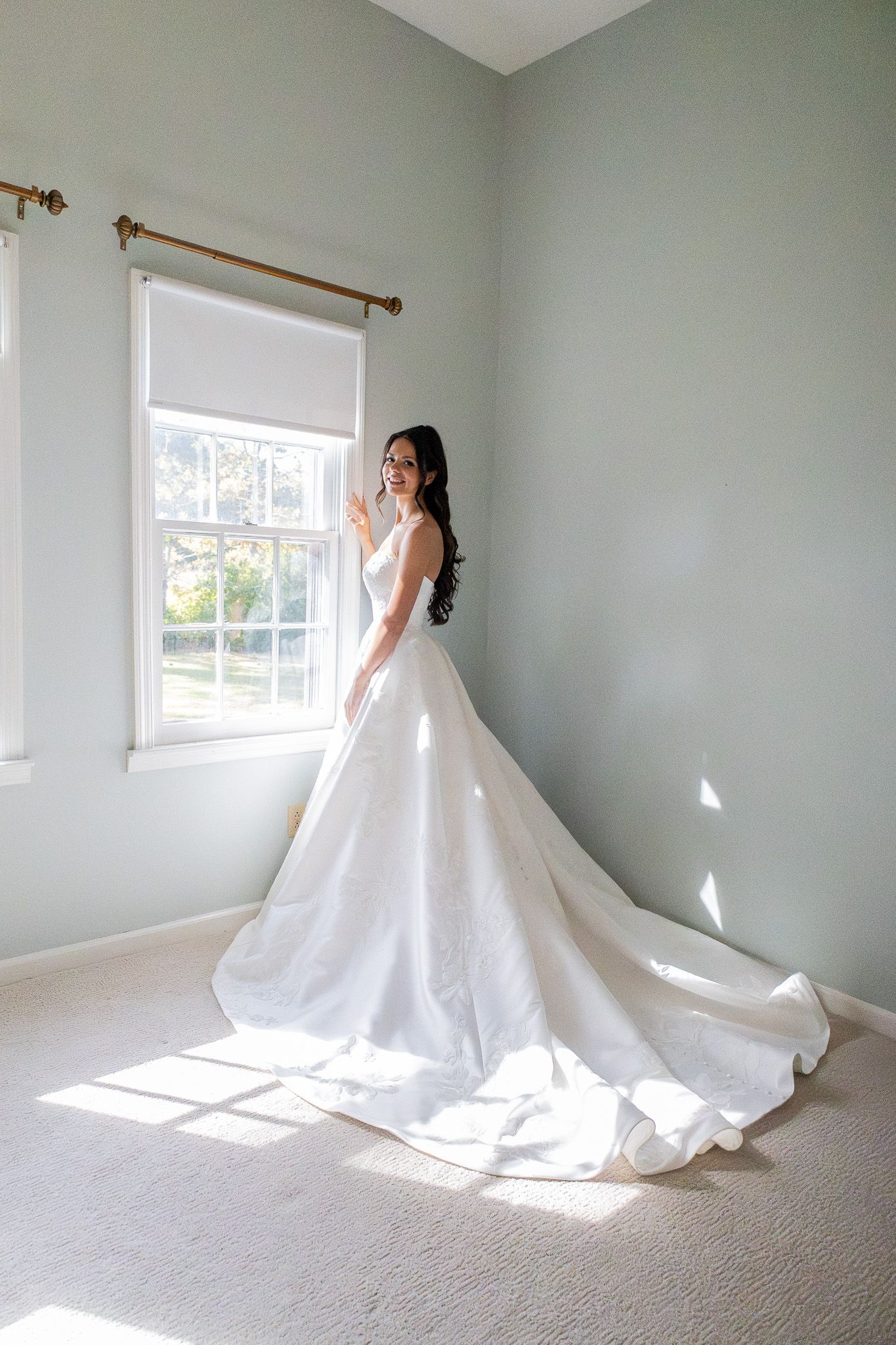 Bride in elegant white ball gown with long train standing by sunlit window in soft gray room.