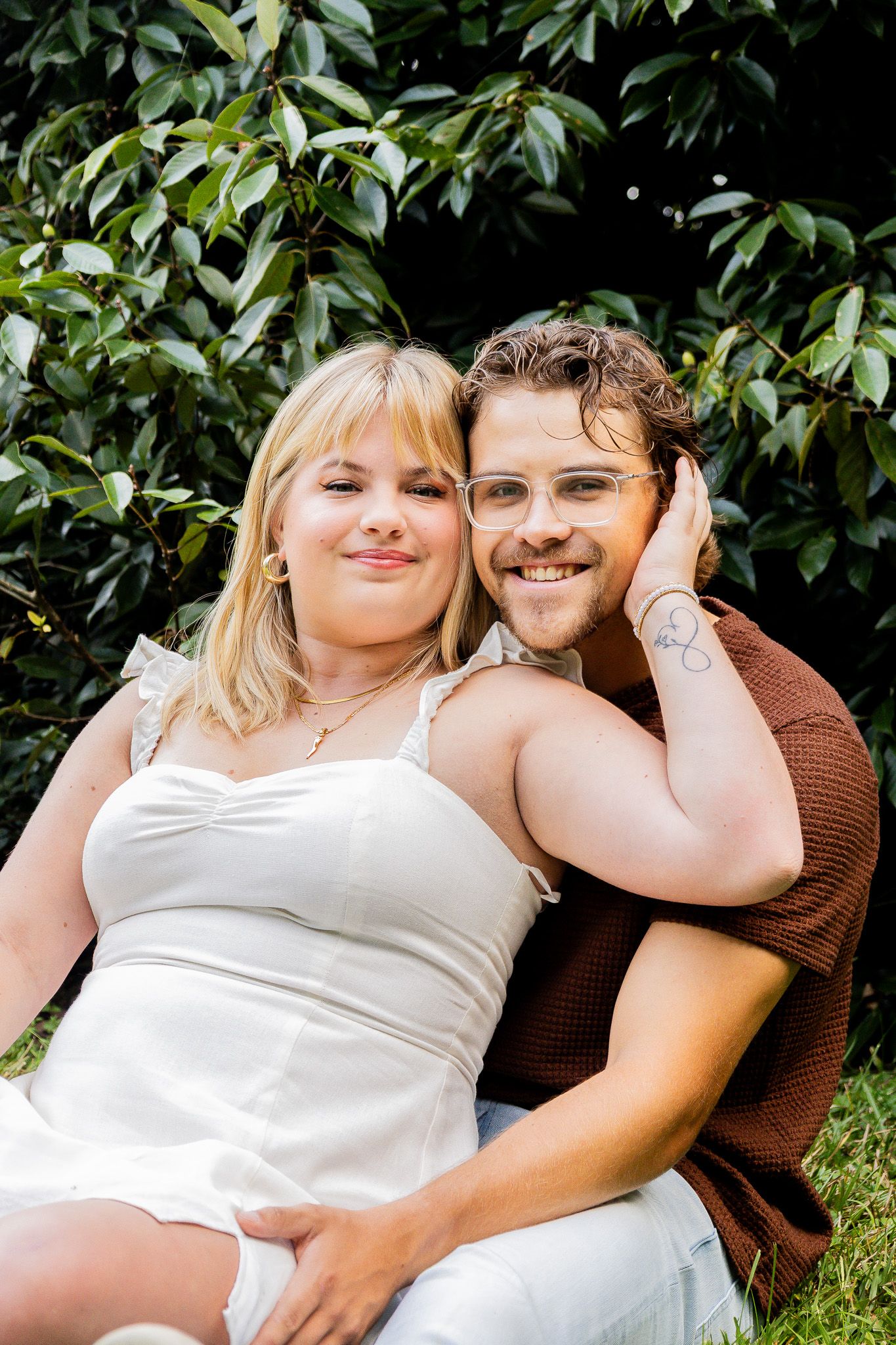 A smiling couple sits together outdoors against lush green foliage, woman in white dress, man in brown shirt wearing glasses.