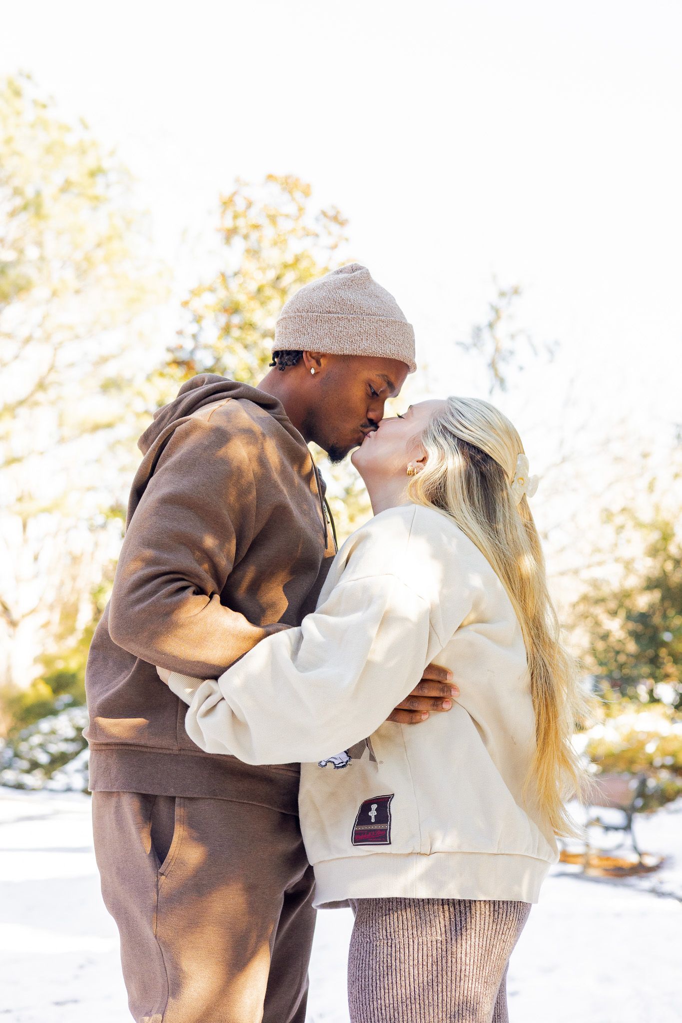 An interracial couple sharing a romantic kiss outdoors in a sunny snowy park, dressed in casual winter attire.