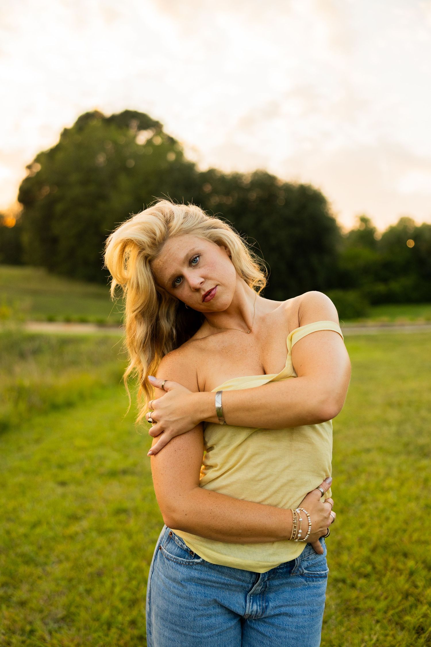 Blonde woman in yellow top and jeans hugging herself in a green field at sunset.