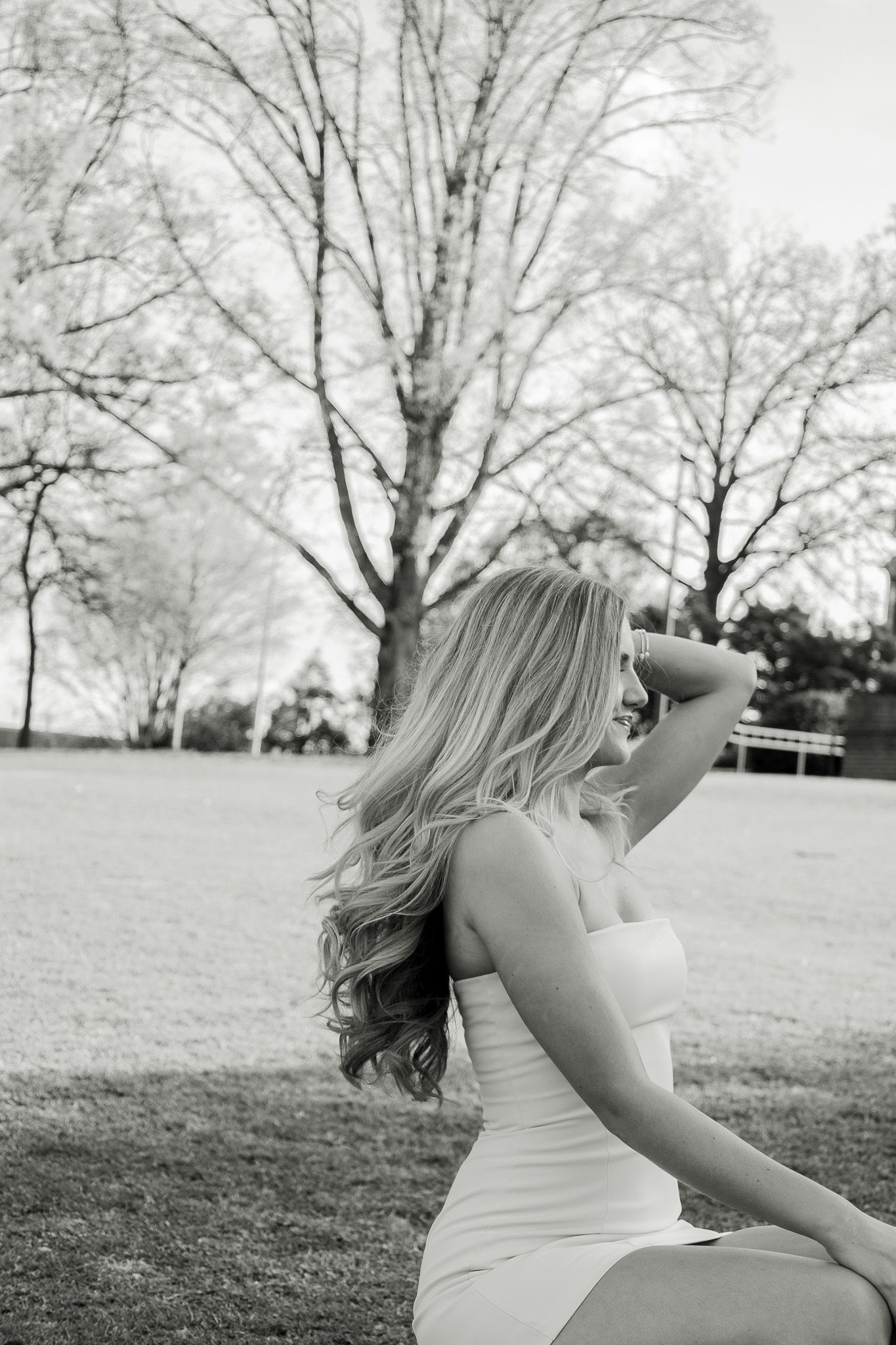 Black and white photo of a blonde woman in a white dress sitting in a park with bare trees in the background.