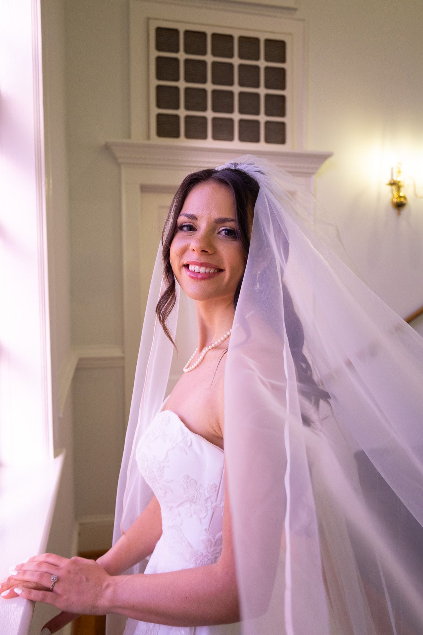 Elegant bride in white lace gown and flowing veil standing on staircase by window in classic interior.