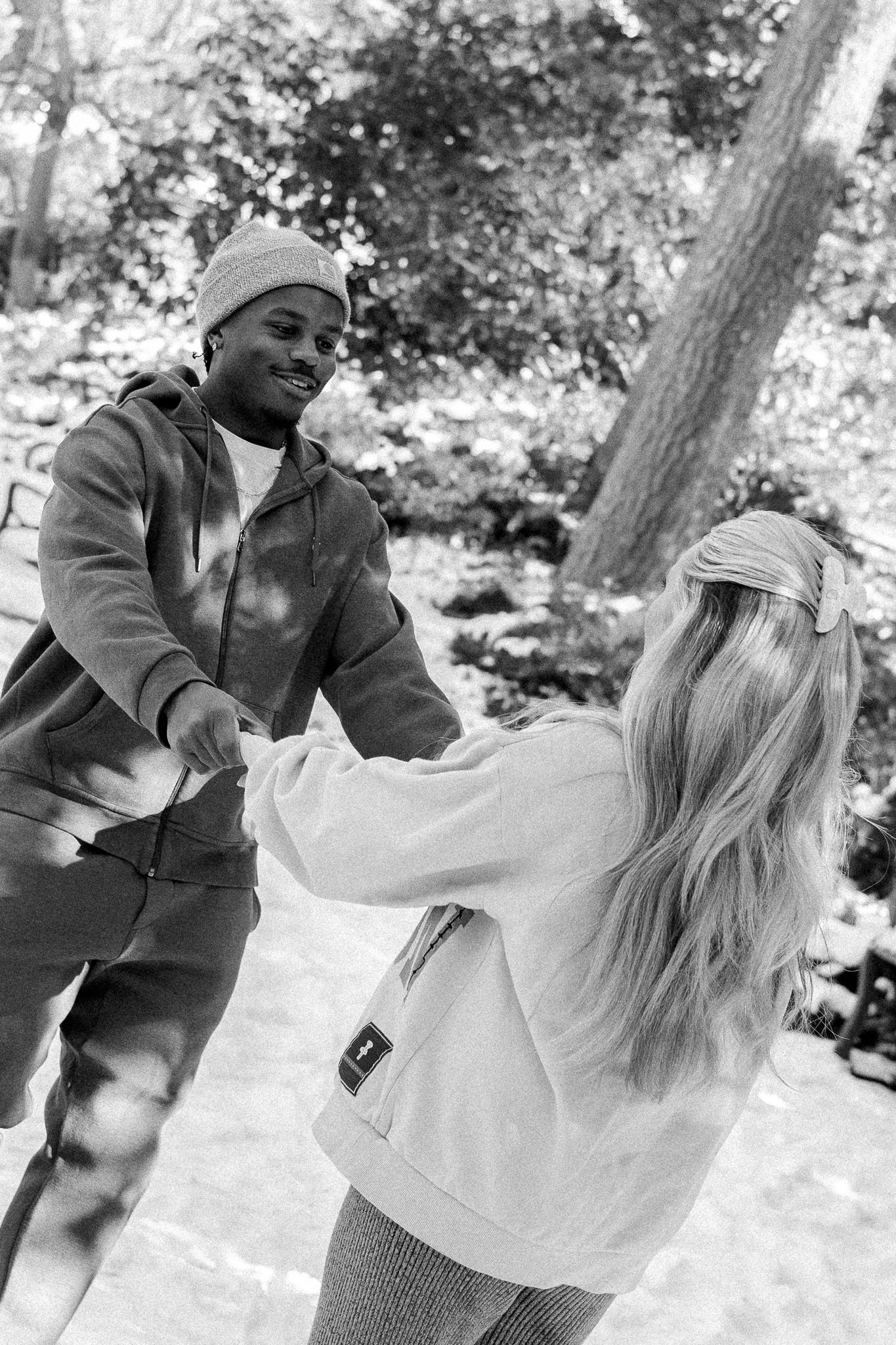 Black and white photo of an interracial couple holding hands and dancing outdoors in a snowy park setting.