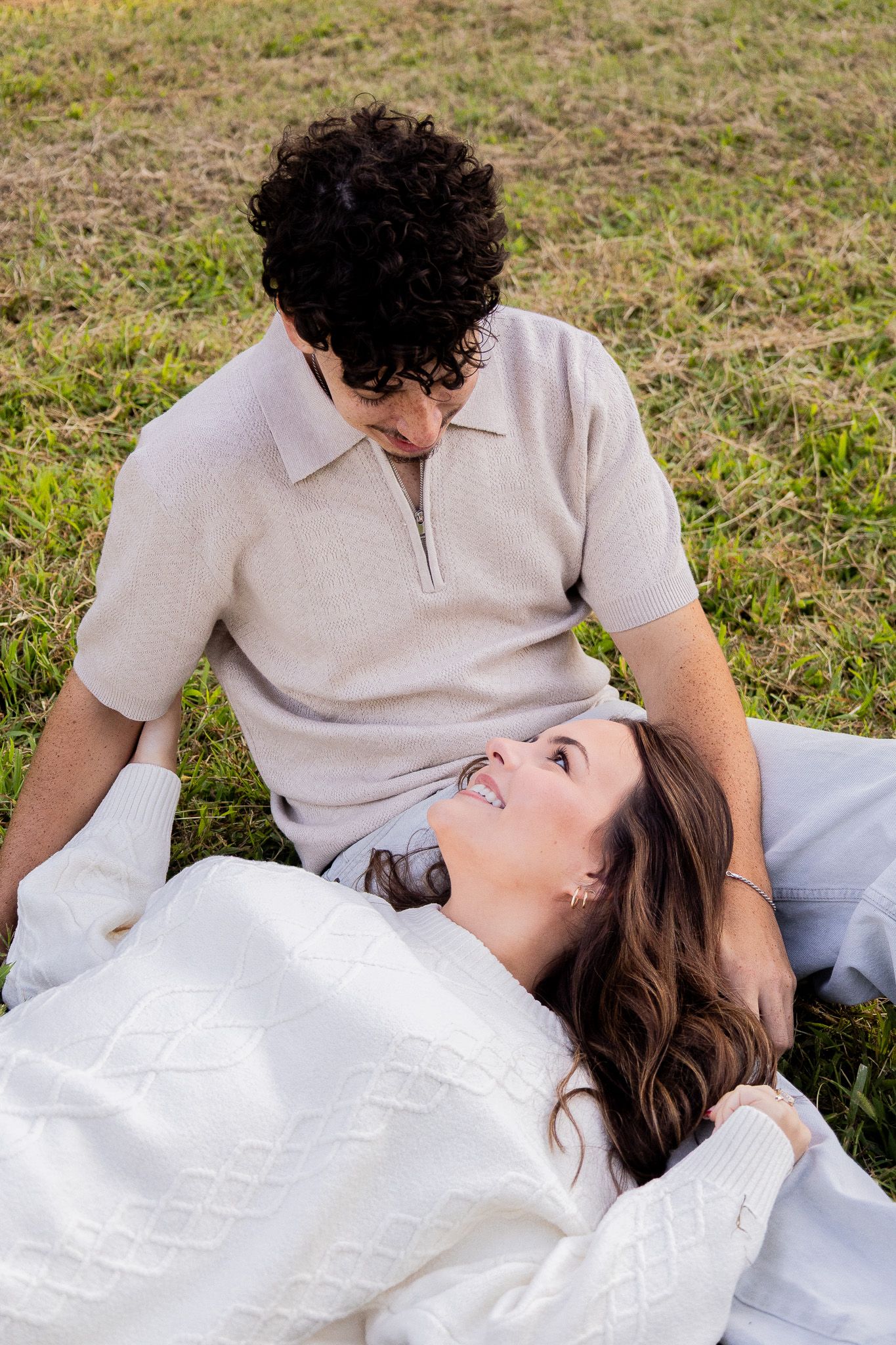 A couple relaxes on green grass, woman lying down smiling up at man in beige polo, both wearing cozy neutral sweaters.
