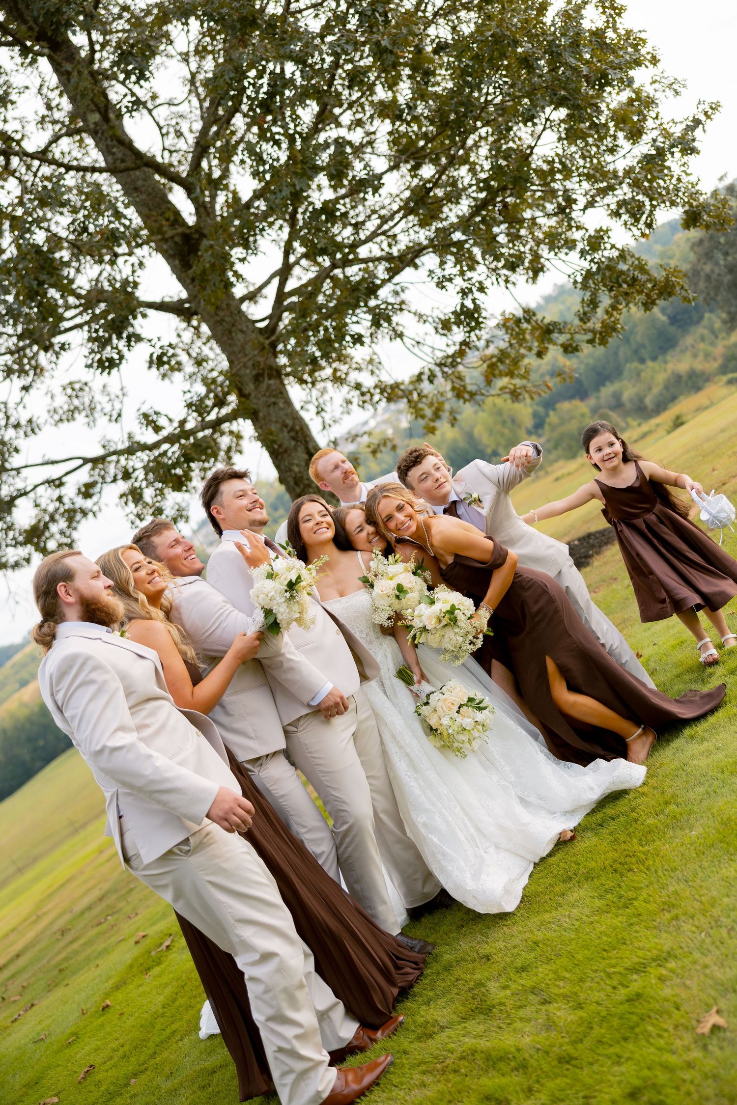 Wedding party posing joyfully outdoors under a large tree on a green lawn with mountains in the background.