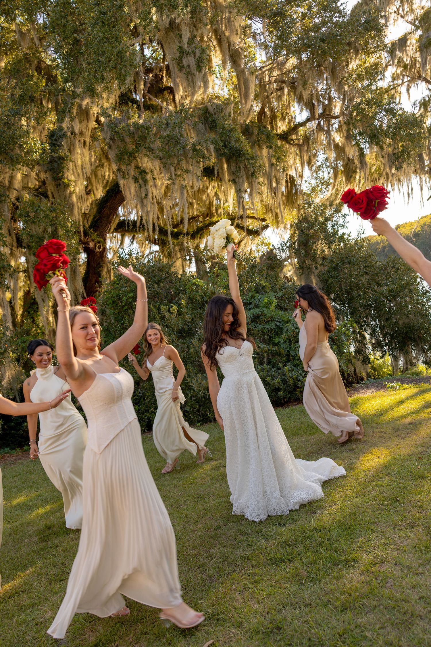 Bride and bridesmaids in white and champagne gowns celebrate outdoors, raising red bouquets under moss-draped oak trees.