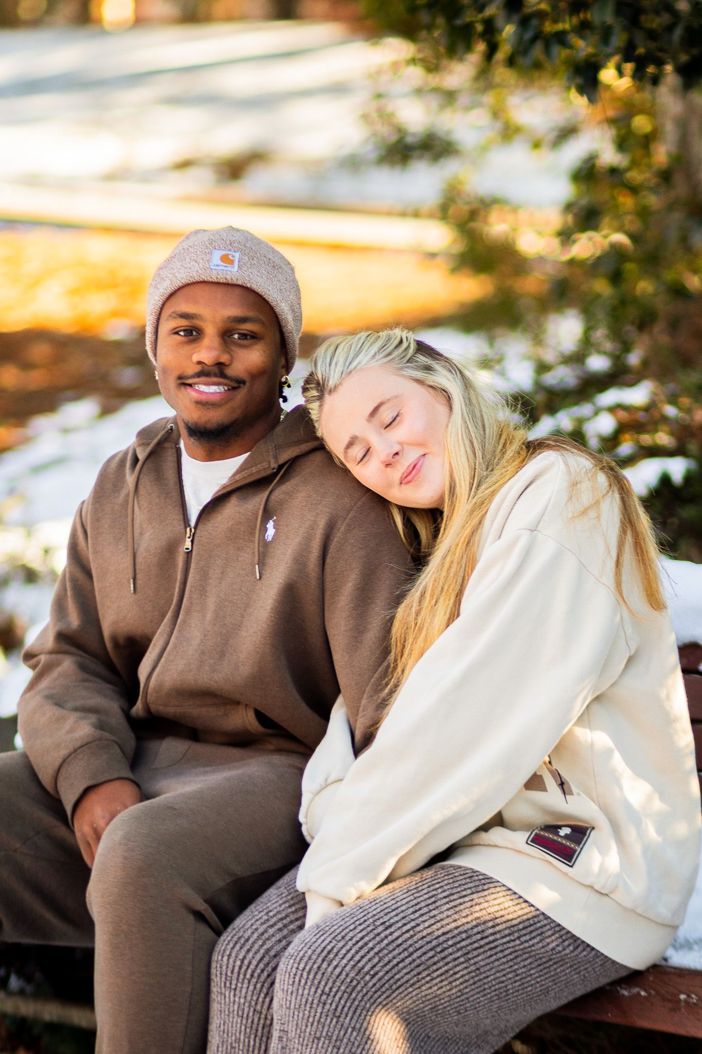 Interracial couple sitting on a bench in winter, woman resting head on man's shoulder, both wearing cozy sweaters.