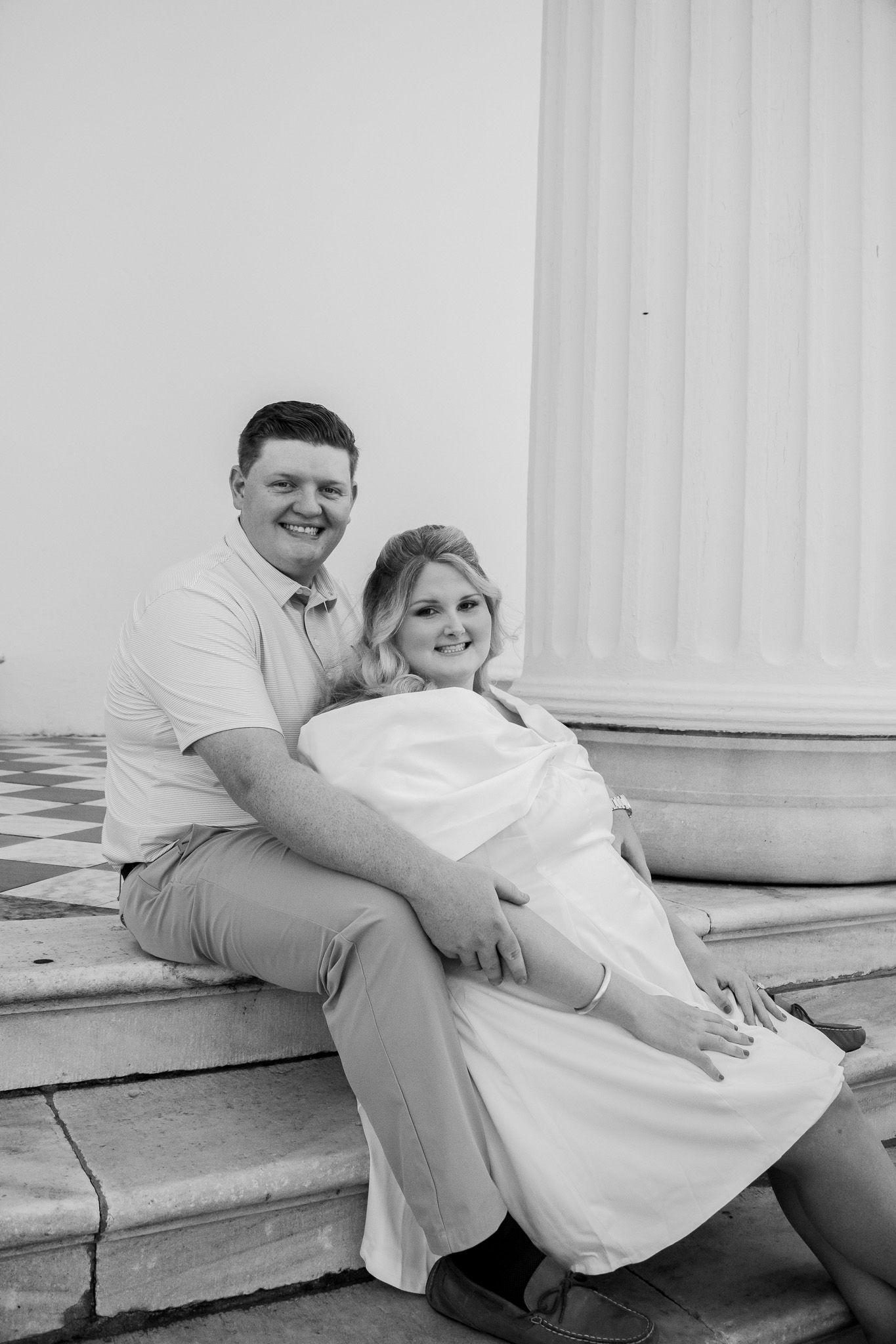 Black and white photo of a smiling couple sitting on steps near a column, woman in white dress, man in polo shirt.