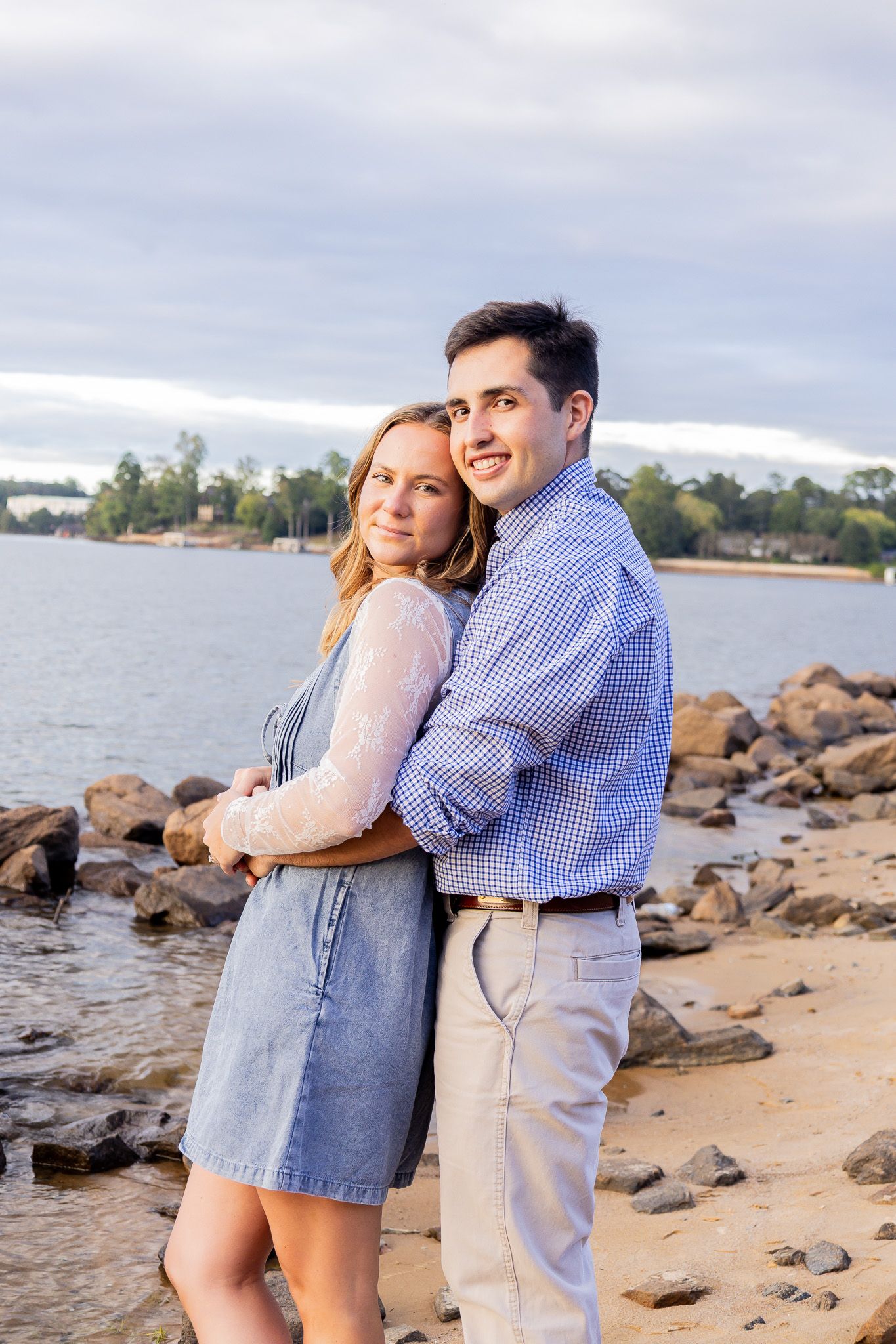 A couple embraces on a rocky beach shore, the woman in a blue dress leaning back into the man in a plaid shirt.