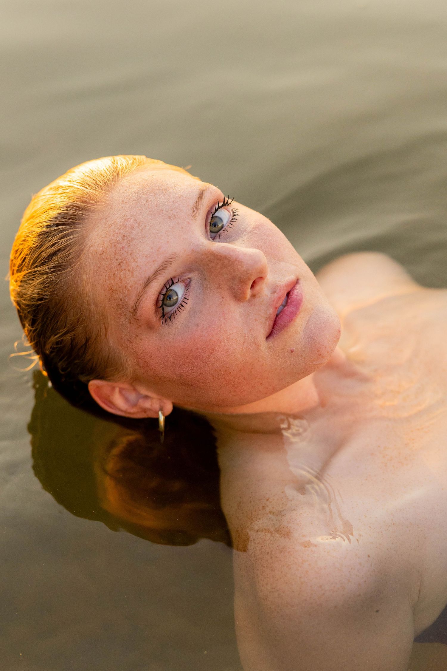 Young redhead woman with freckles and blue eyes submerged in calm water, looking up with a serene expression.