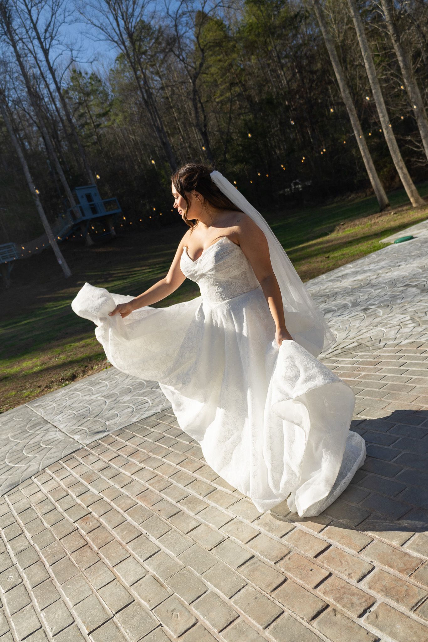 Bride in white strapless gown and veil twirling on brick patio outdoors with trees in background.