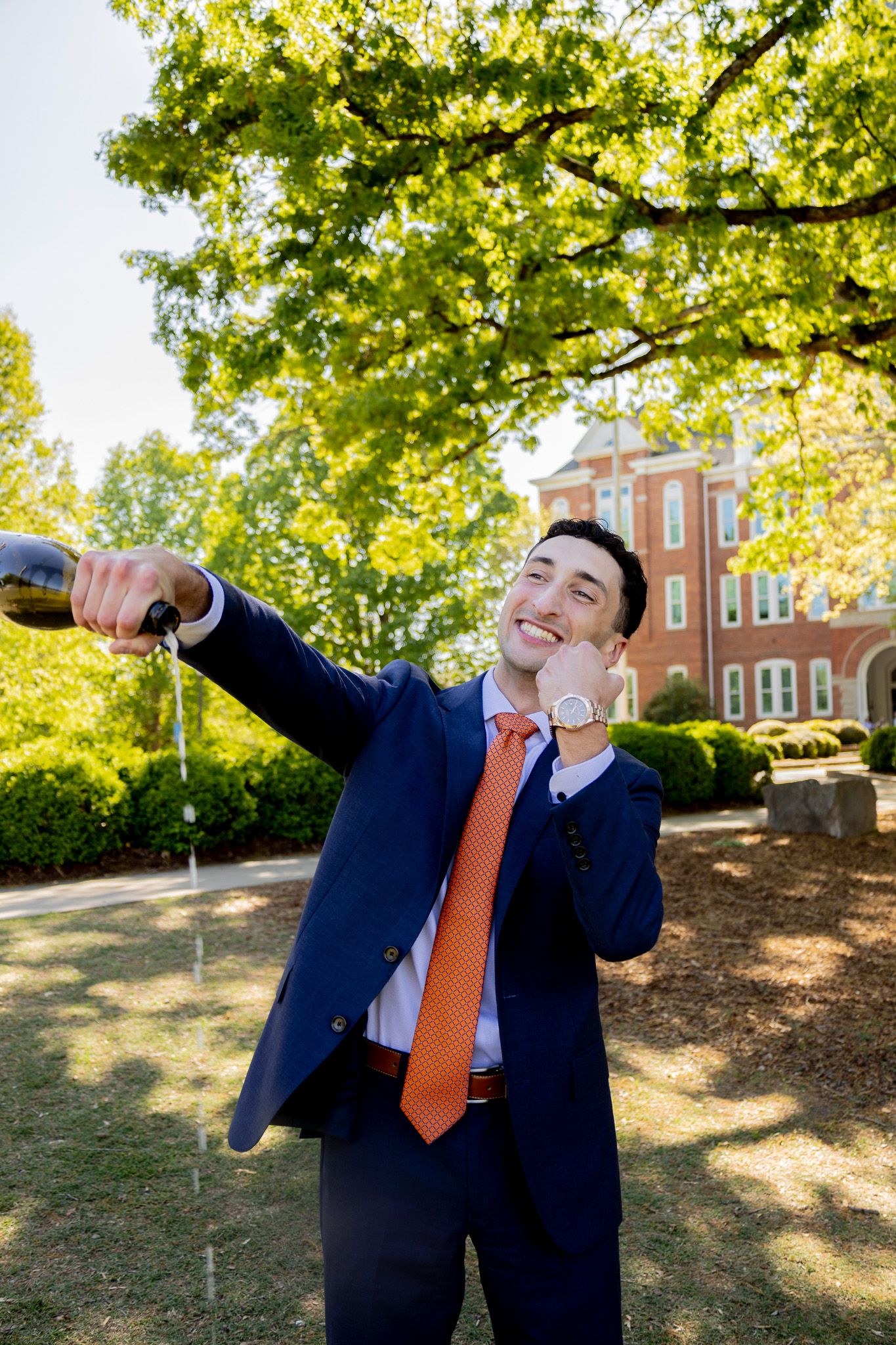 Man in navy suit and orange tie celebrating outdoors, pouring champagne while smiling near a brick building.