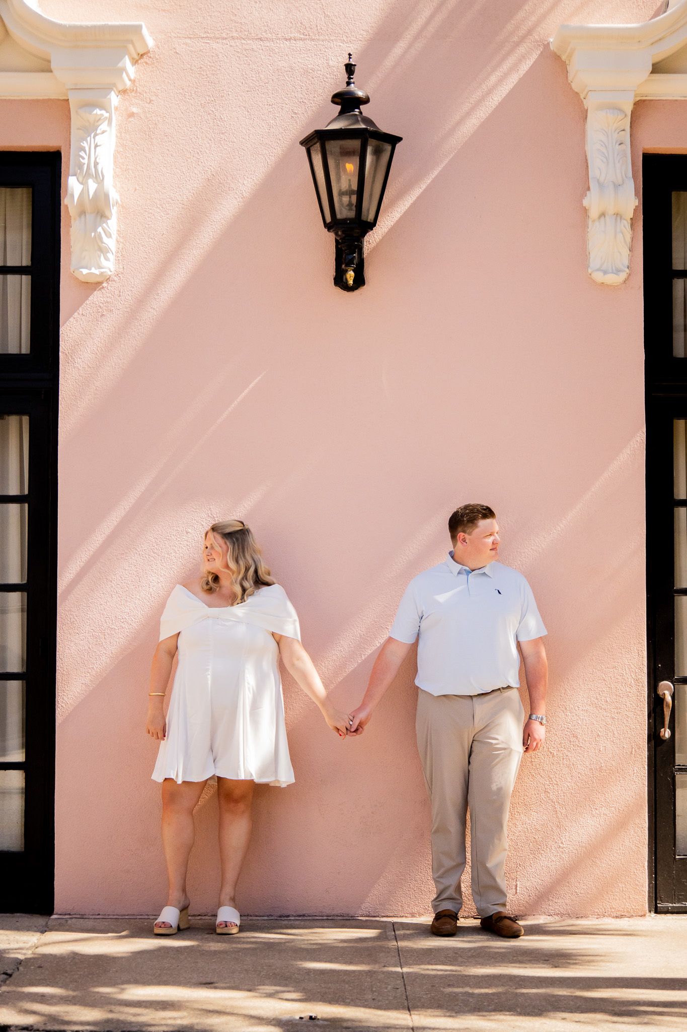 Couple holding hands in front of a pink stucco wall with a black lantern light fixture between them.
