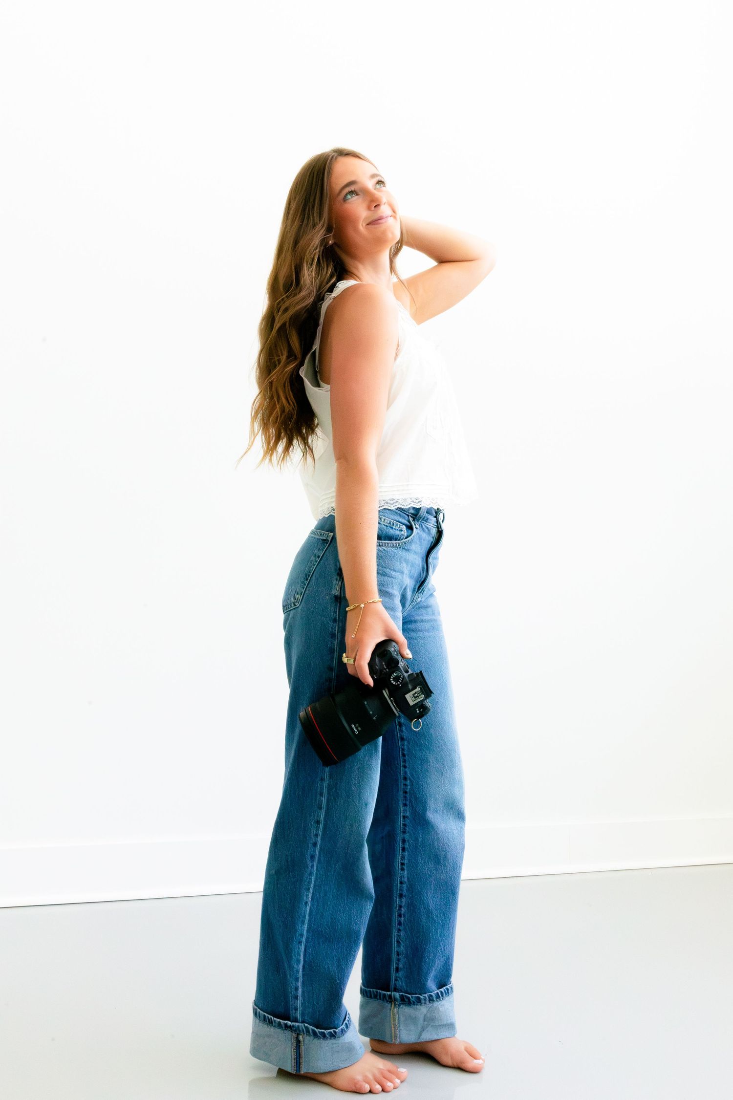 Woman in wide-leg jeans and white tank top holds camera, posing barefoot against white background.