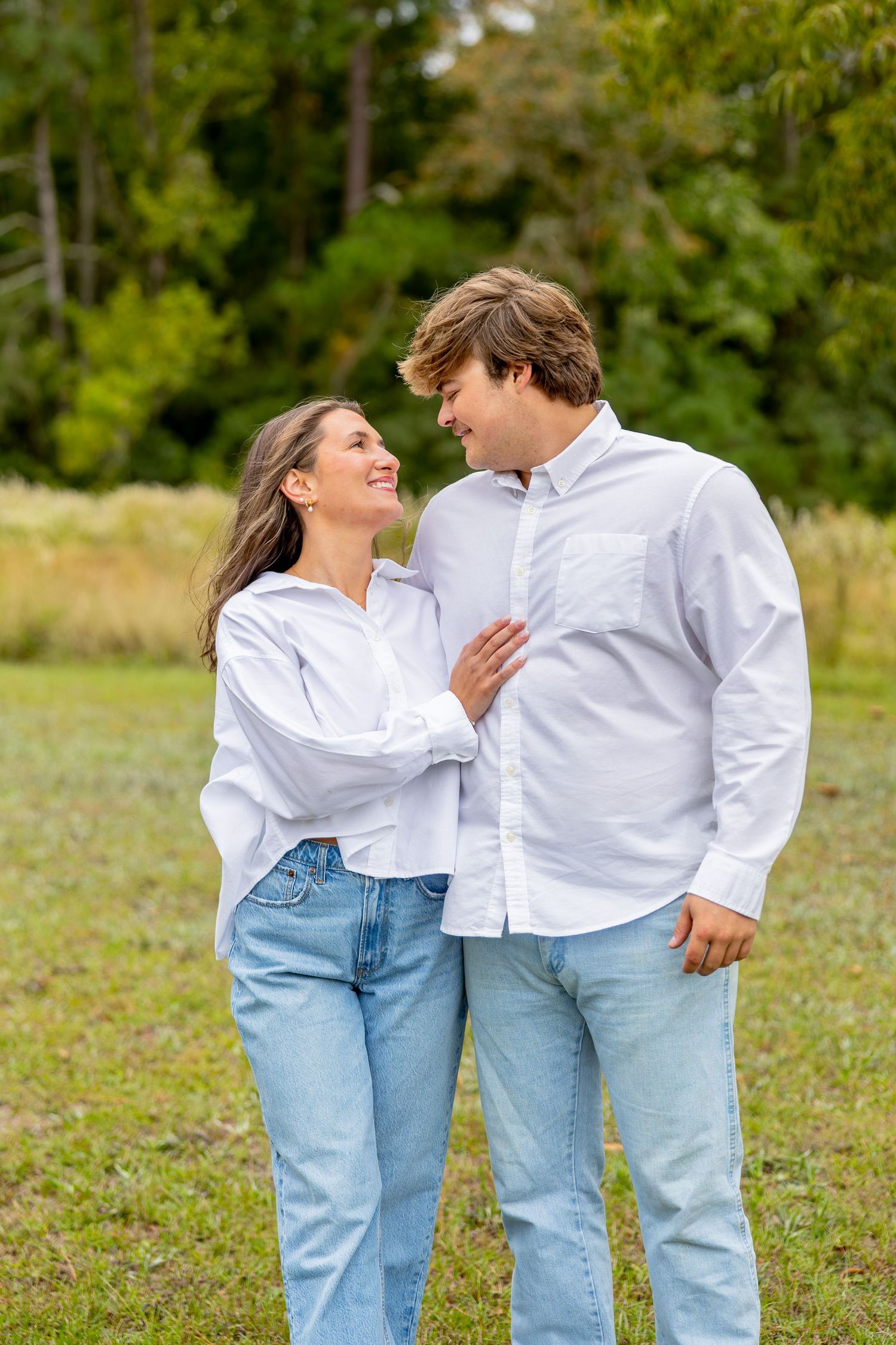 A smiling couple in matching white button-up shirts and light-wash jeans embrace while standing in a grassy field with trees behind them.