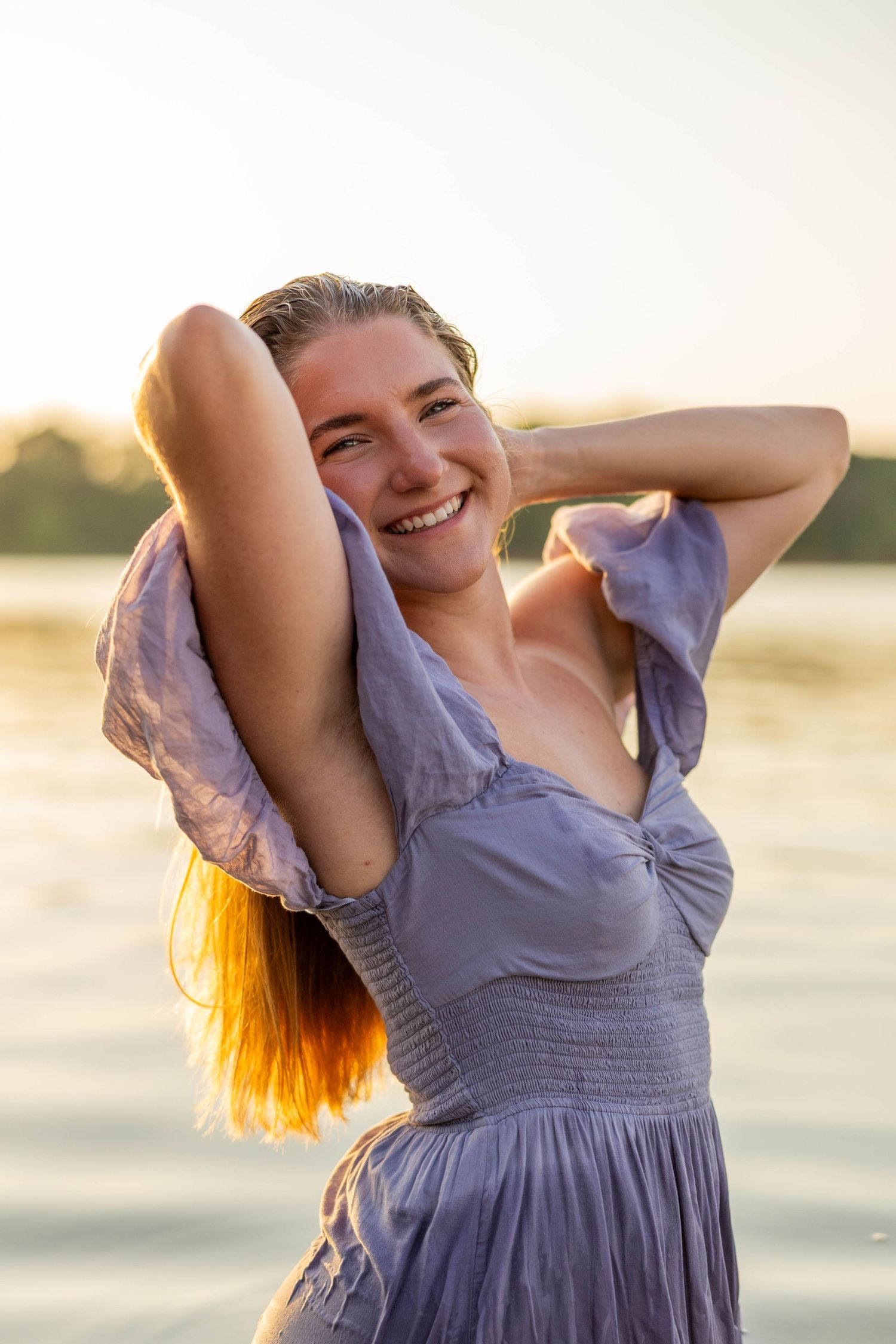 Young woman in lavender dress smiling joyfully by a lake at golden hour sunset, hands behind her head.