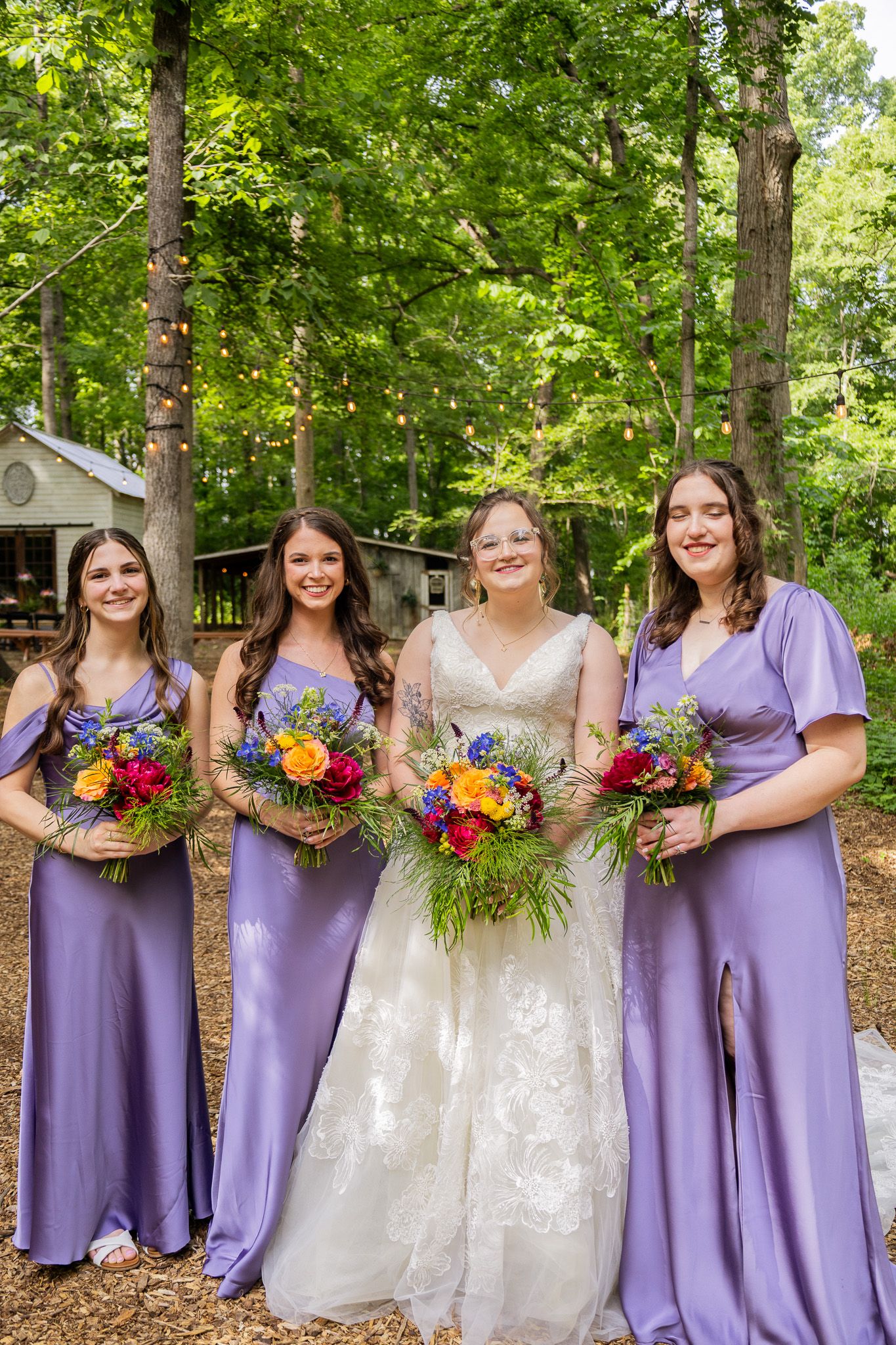 Bride in white lace gown with three bridesmaids in purple dresses holding colorful bouquets outdoors.
