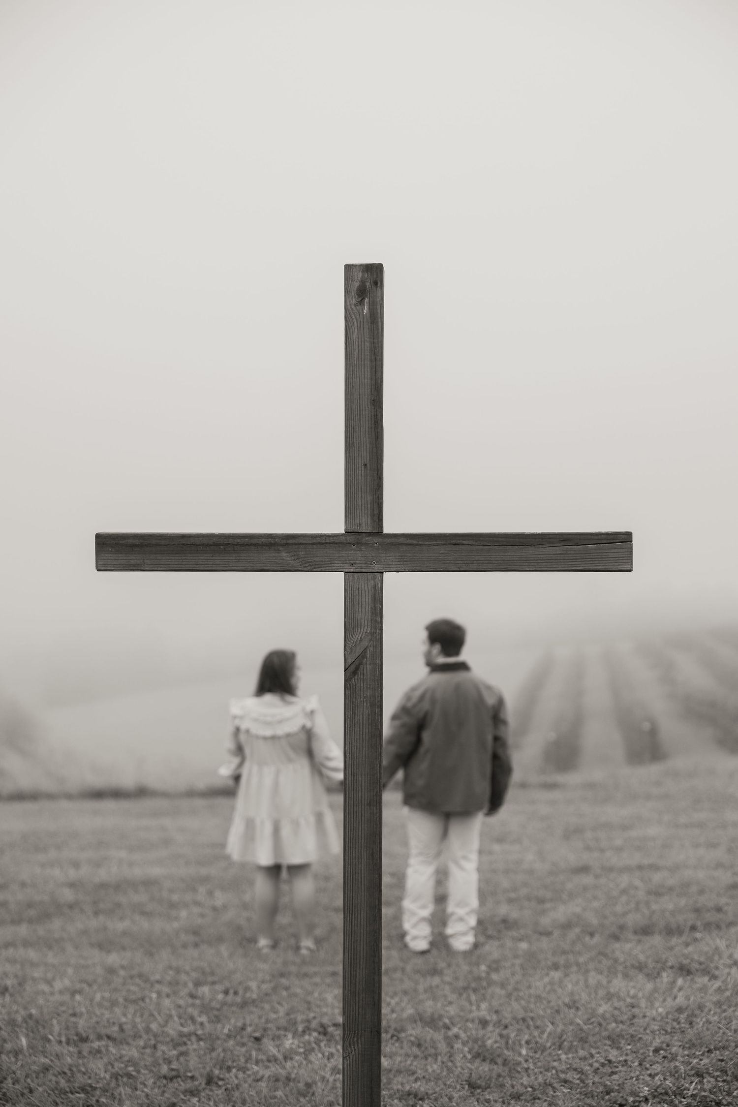 Black and white photo of a couple holding hands behind a wooden cross in a misty field.