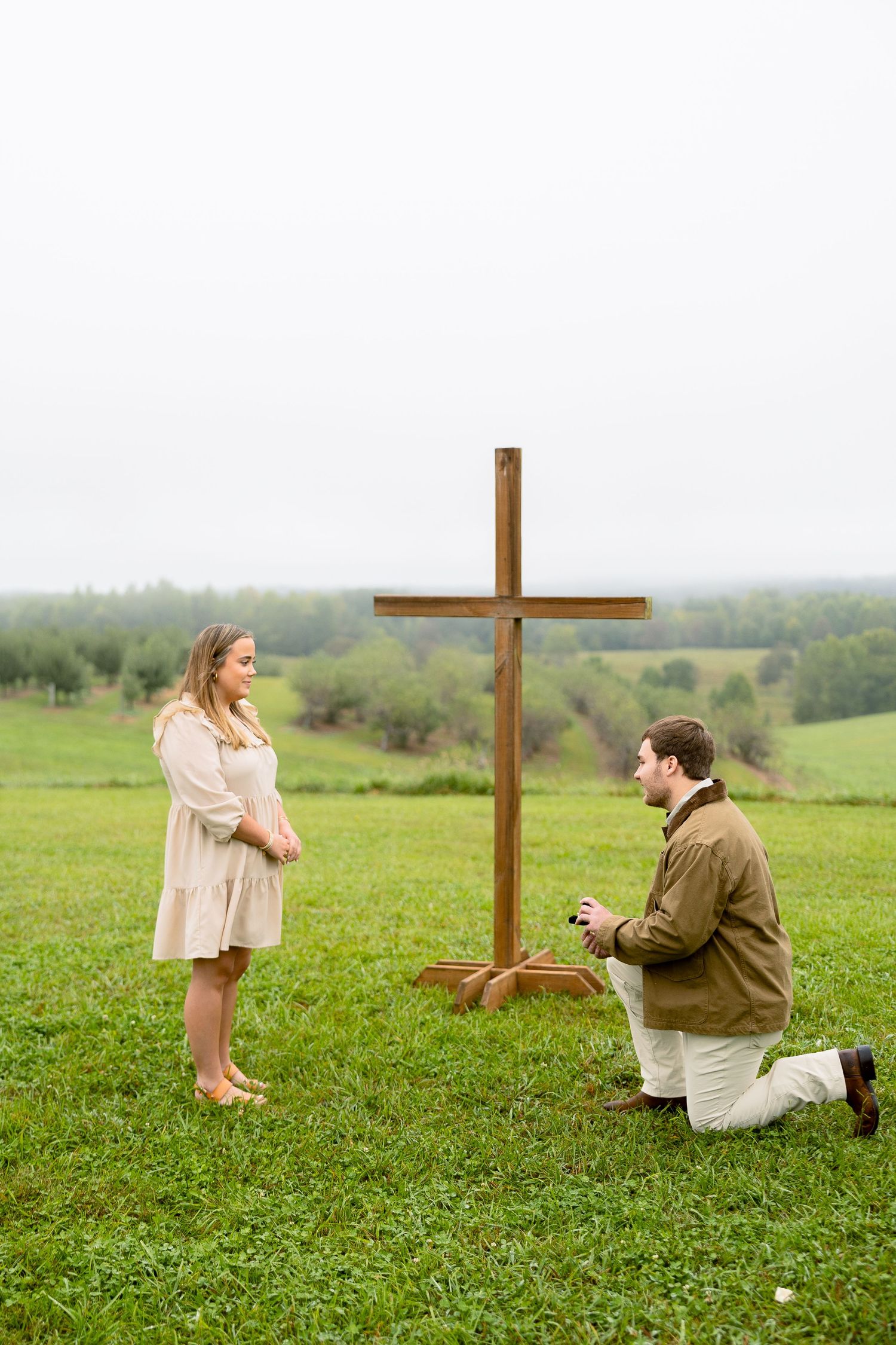 Man proposes on one knee to woman in field with wooden cross backdrop on overcast day.