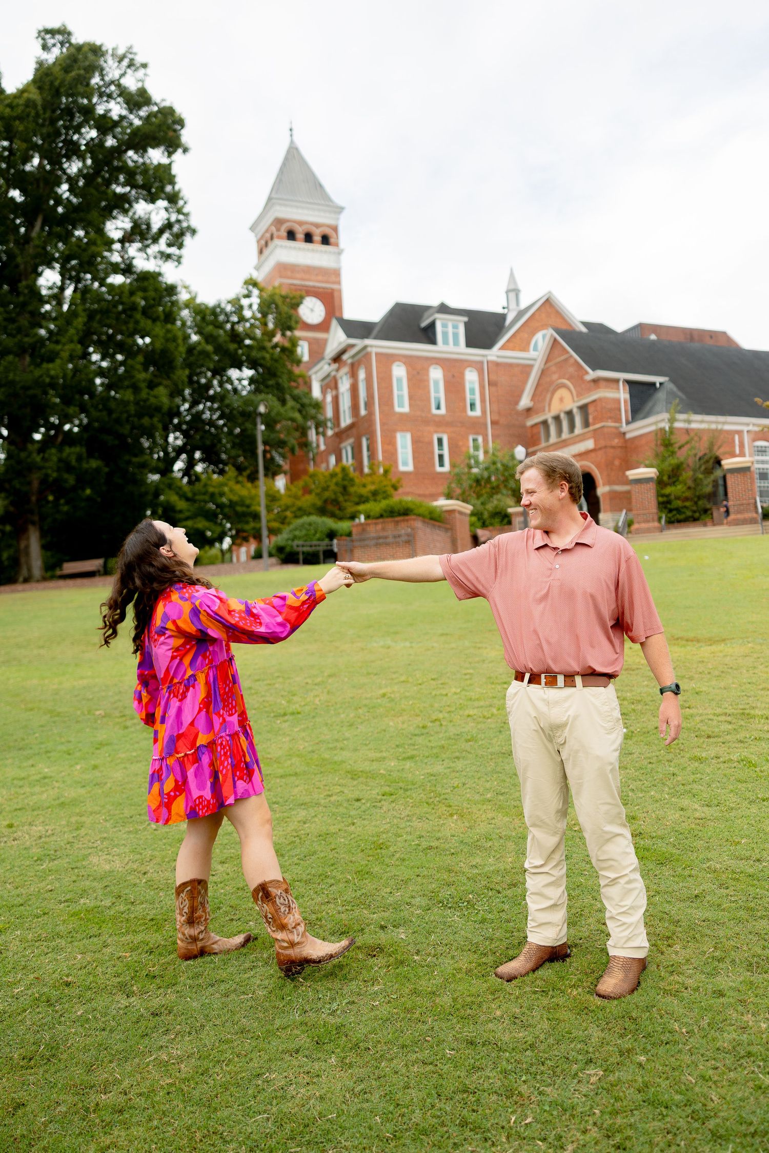 A couple dances on a green lawn in front of a red brick building with a white steeple on a cloudy day.