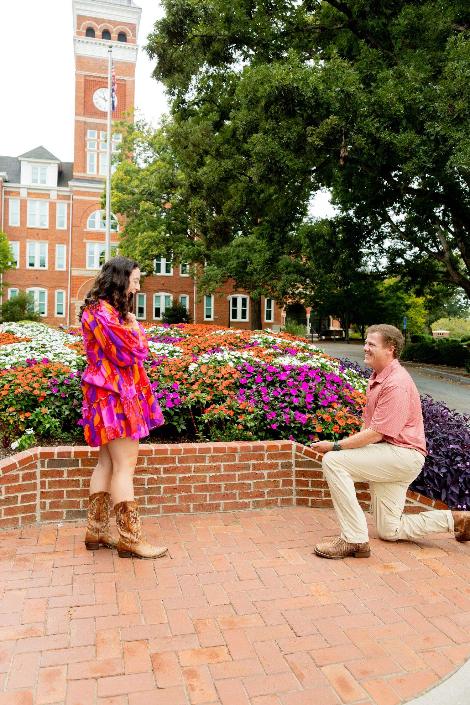 Man proposes on one knee to woman in pink dress near colorful flower garden at brick university campus.