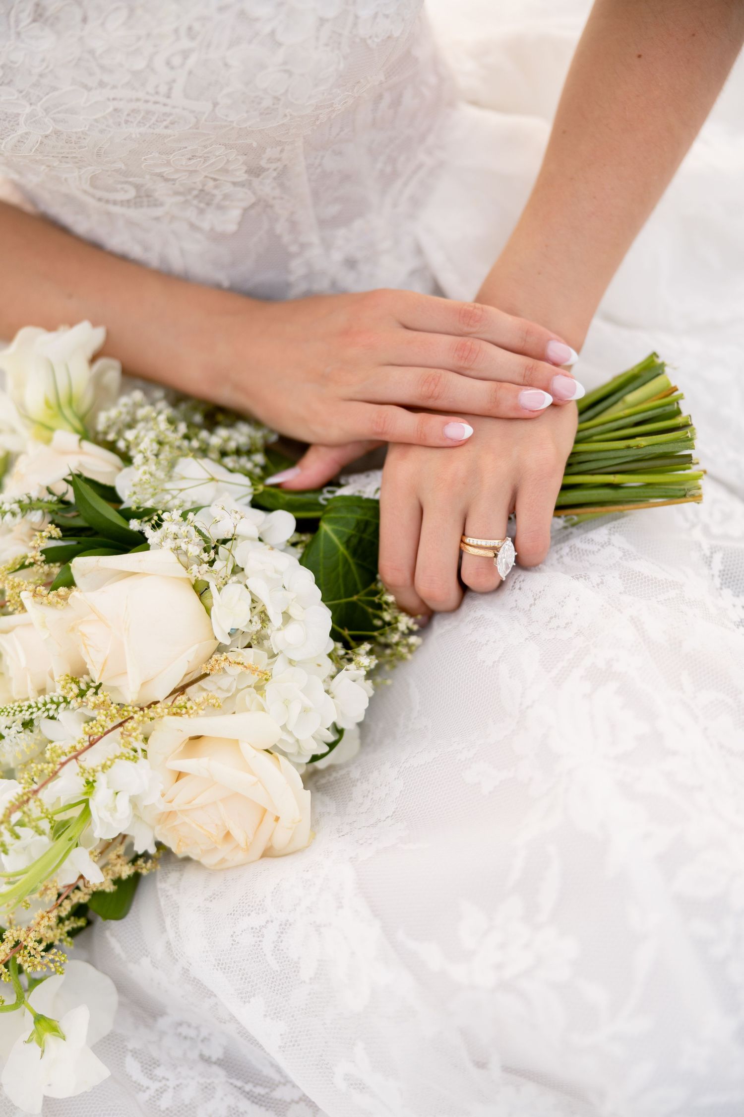 Bride in white lace dress holding elegant bouquet of cream roses with gold accents, showcasing diamond engagement ring.