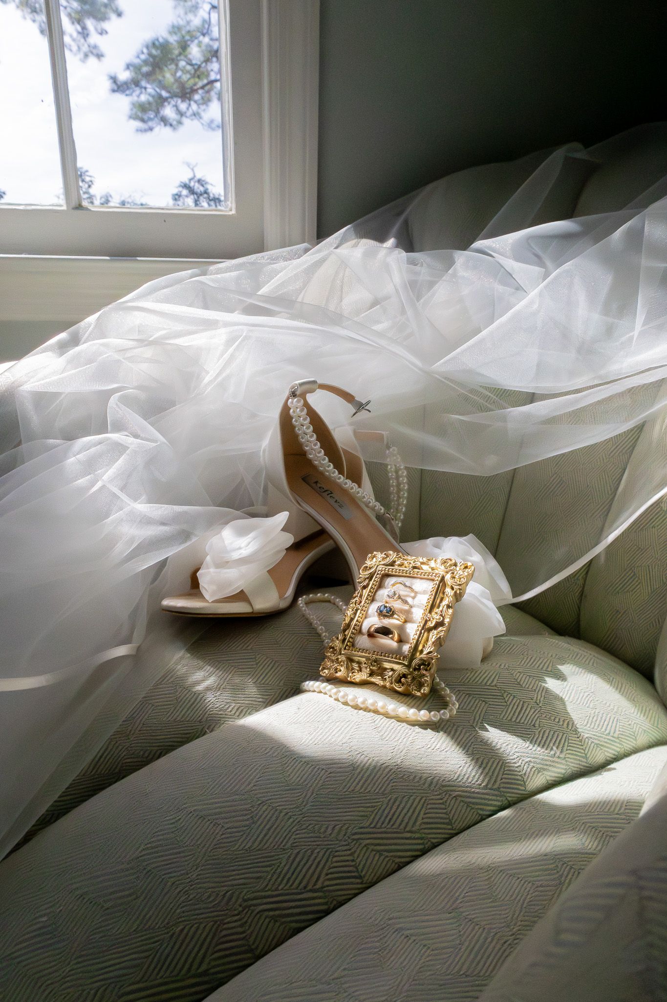 Bridal accessories arranged on a chair, featuring ivory heels with pearl straps, a gold ornate purse, and a flowing white tulle veil.