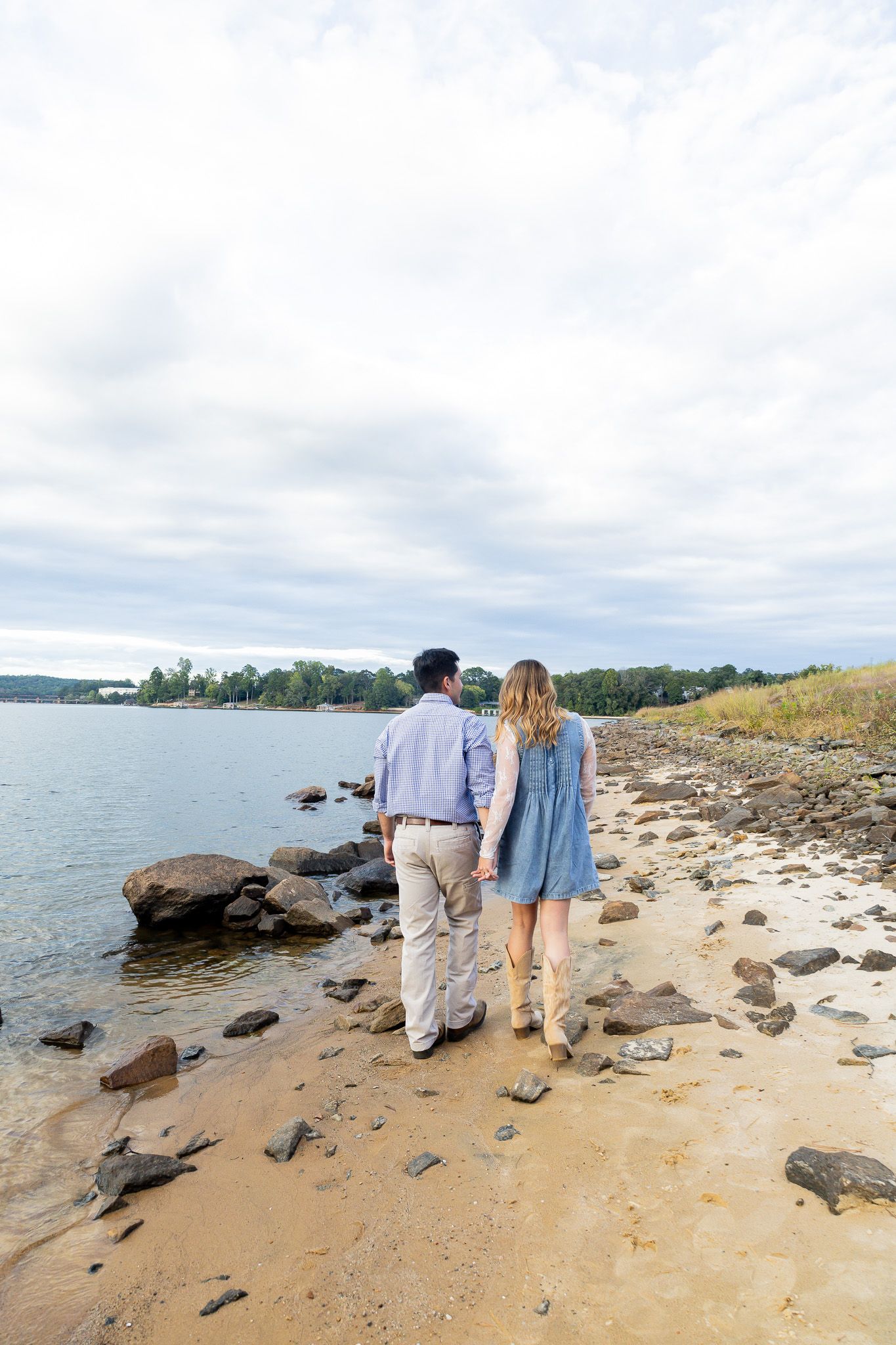 Couple holding hands on rocky shoreline, viewed from behind, gazing at calm lake with tree-lined horizon under cloudy sky.